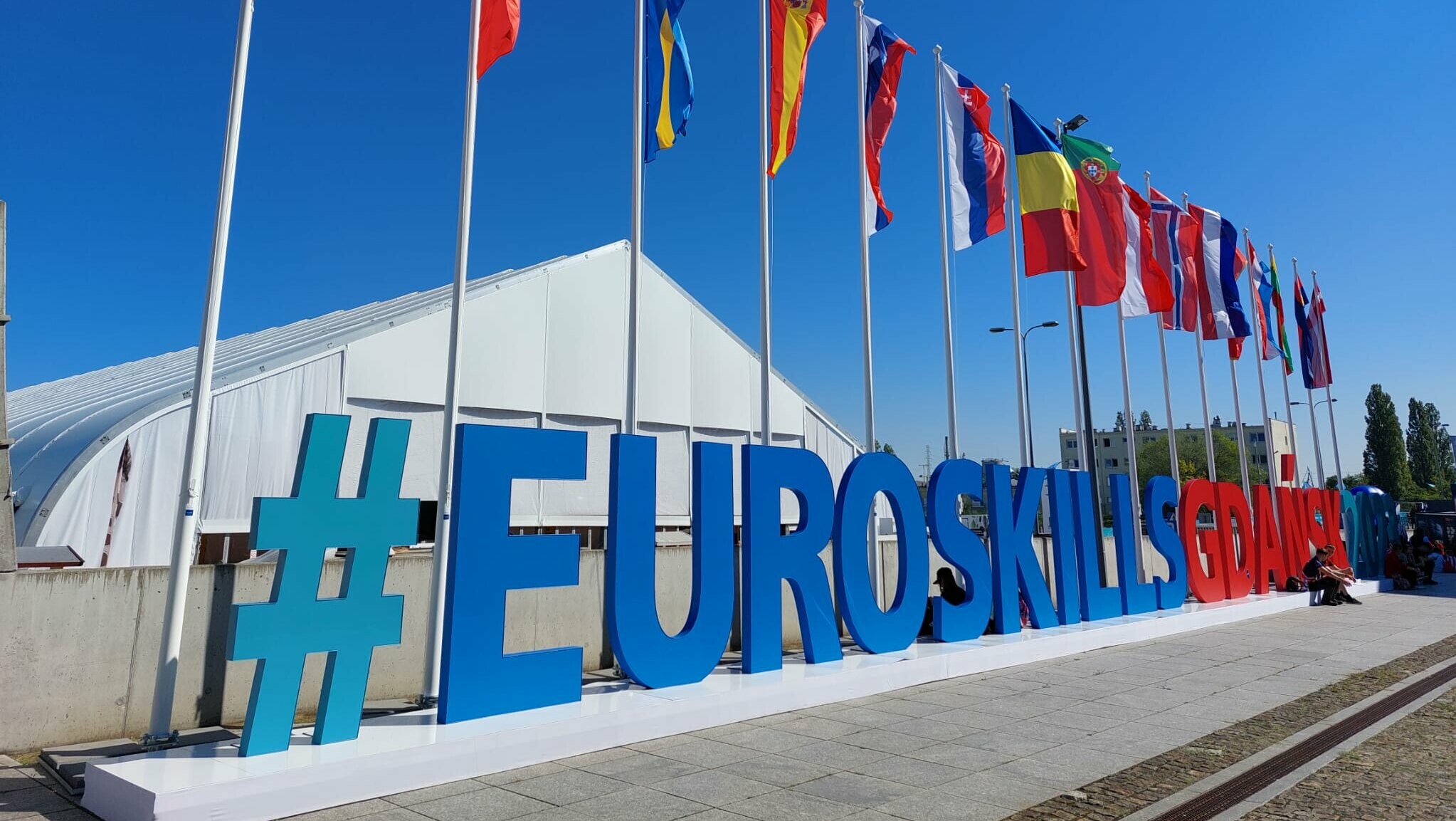 Vor einem Eventzelt sind die Länderflaggen sowie die Aufschrift #EuroSkillsGdansk2023 zu sehen.
