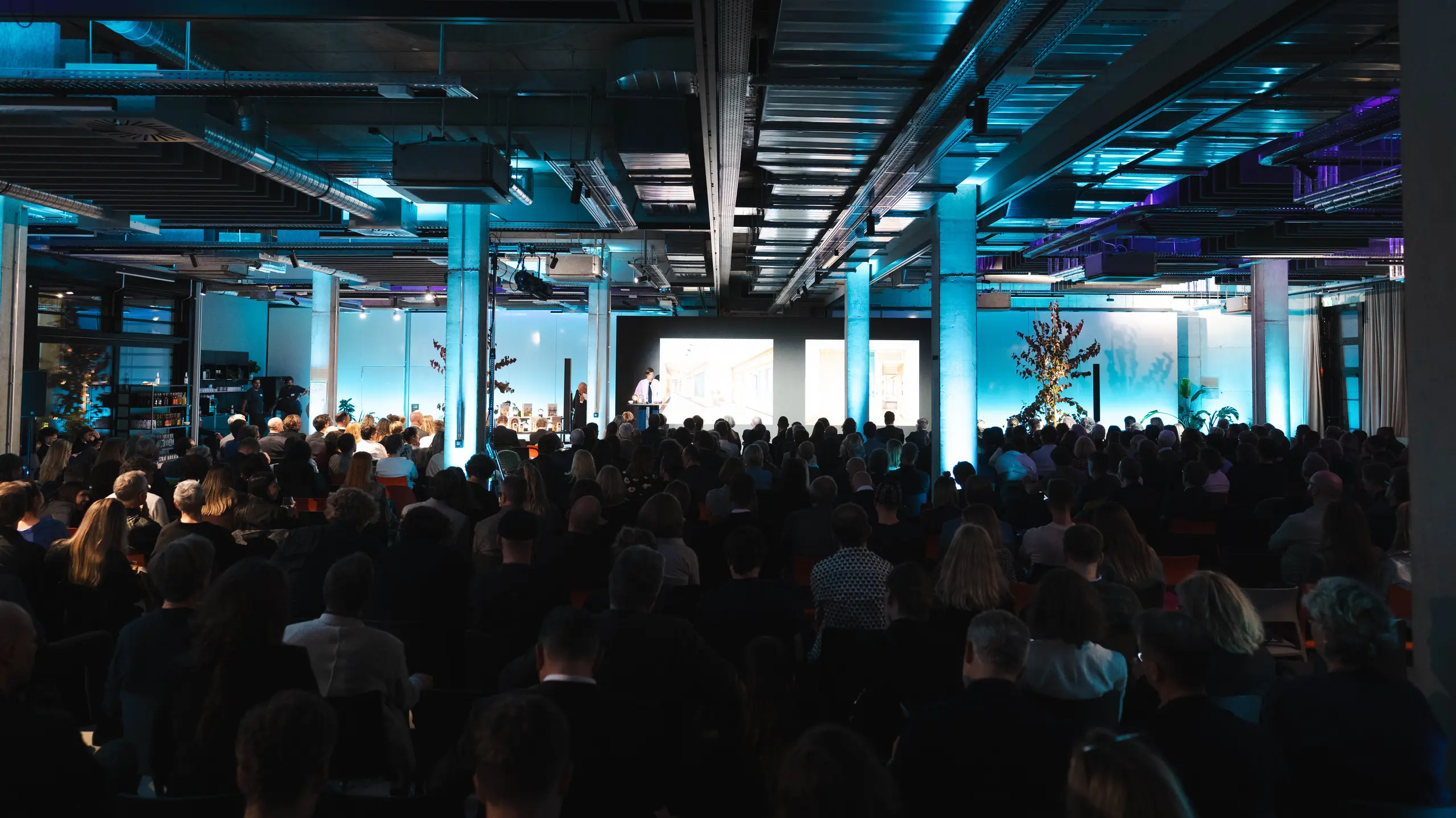 Eine stimmungsvolle Szene von der Preisverleihung „Wohngebäude des Jahres 2025“: Blick auf einen vollbesetzten Saal in einer modernen Industriehalle mit auffälliger blauer Beleuchtung. Im Vordergrund zeigt eine Bühne mit großem Bildschirm die Präsentationen. Die Architektur des Raumes mit ihren freiliegenden Stahlträgern und Betonpfeilern unterstreicht den innovativen Charakter der Veranstaltung, bei der nachhaltige Lösungen wie das PREFALZ-Solarmodul gefeiert werden.