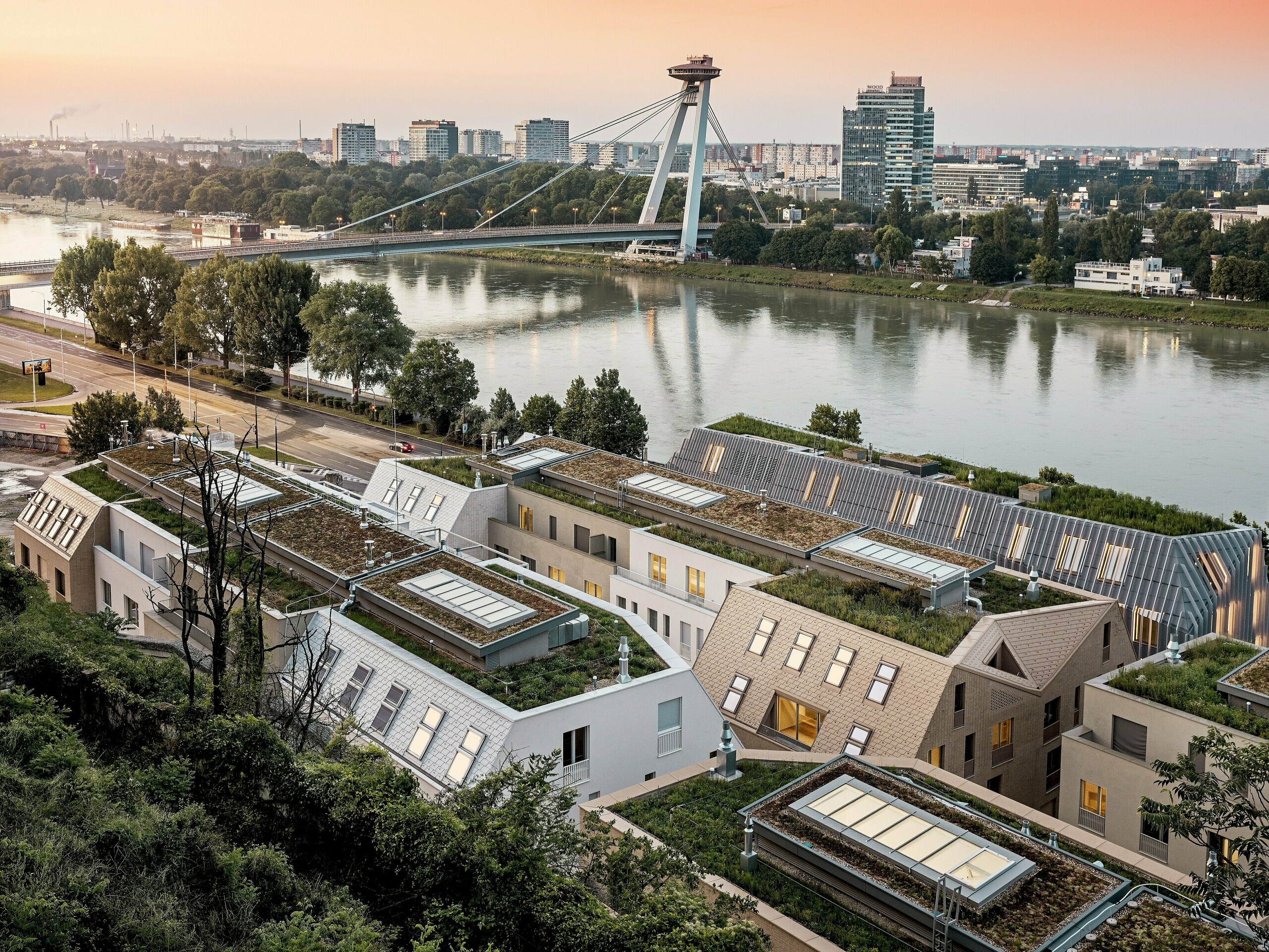 Spektakulärer Blick über das Stadtquartier Vydrica in Bratislava mit Donauufer und markanter Brücke im Abendlicht: Die vielschichtige Dachlandschaft mit begrünten Flächen, präzise integrierten Dachfenstern und steilen Satteldächern aus PREFA Aluminium Dachschindeln in Prefaweiß, Sandbraun und Bronze wird zum visuellen Highlight. Die klaren Linien und harmonischen Farbtöne verbinden Architektur und Umgebung eindrucksvoll. PREFA Aluminium überzeugt durch extreme Langlebigkeit, geringes Gewicht, hohe Witterungsbeständigkeit und flexible Gestaltung – ideal für komplexe, nachhaltige Dachlösungen.