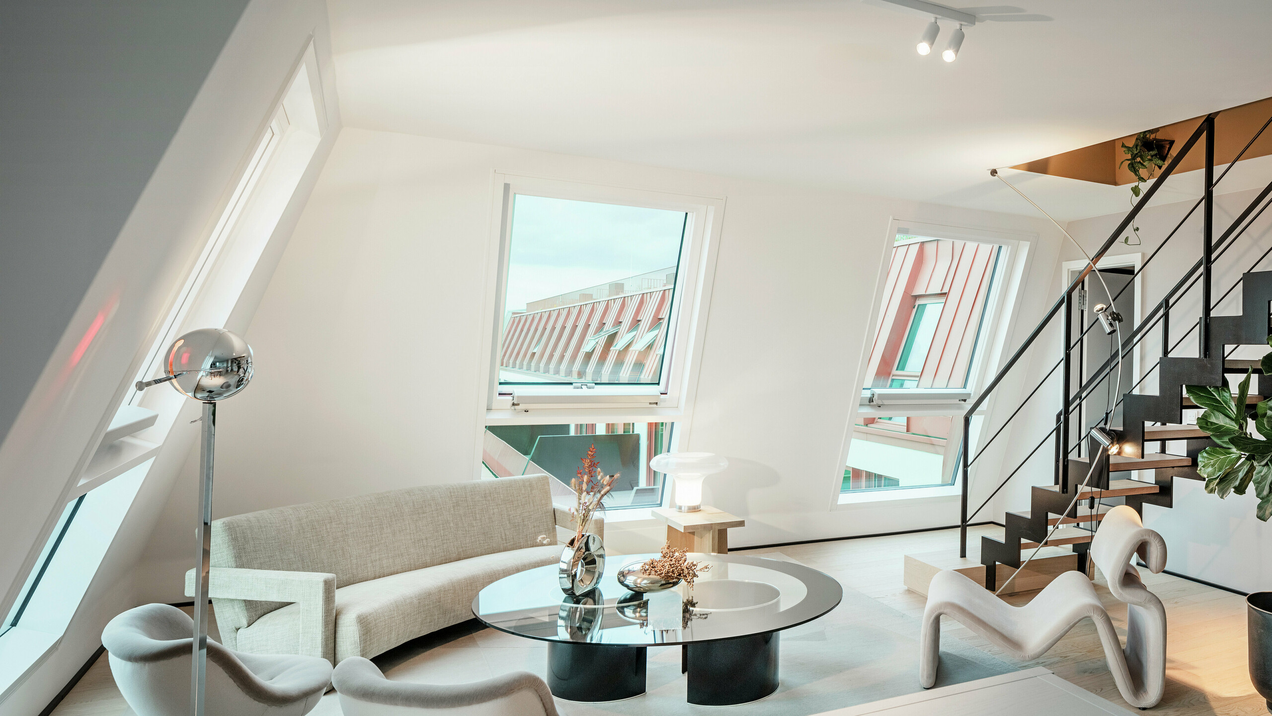 Innenansicht eines lichtdurchfluteten Penthouses im Wohnprojekt Van B in München mit Blick auf das hochwertige PREFALZ Aluminiumdach in der Sonderfarbe IGP Orange HDP. Die großen Fensterflächen lassen viel Tageslicht herein und unterstreichen das moderne Wohnkonzept. Klare Linien, Designermöbel und die präzise Verarbeitung der Dachanschlüsse zeigen, wie hochwertige und stilvolle Architektur nicht nur außen, sondern auch innen umgesetzt werden kann.
