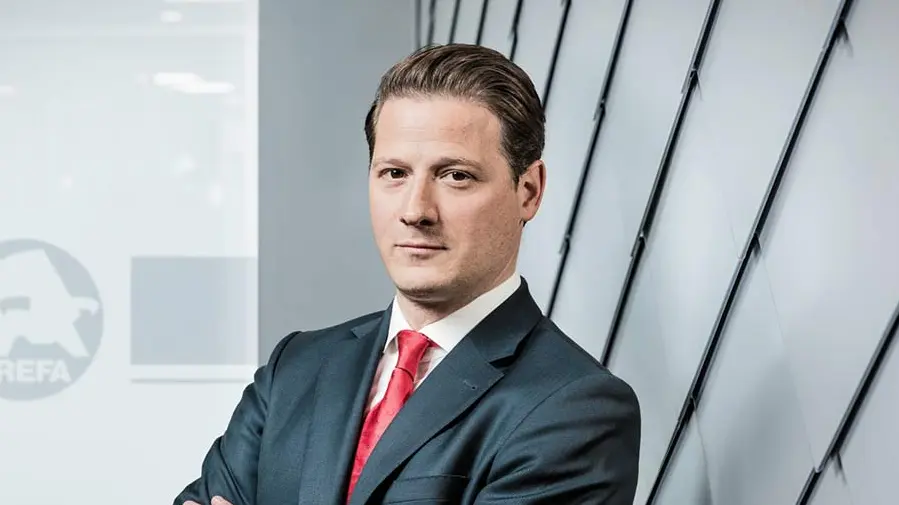 Portrait des neuen PREFA Geschäftsführers Leopold Pasquali, mit dunklem Anzug und roter Krawatte vor PREFA Aluminium Fassade und Glaswand