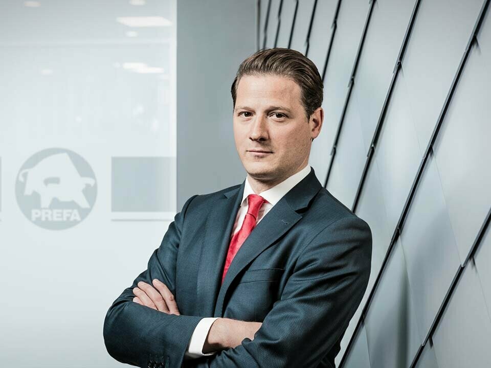 Portrait des neuen PREFA Geschäftsführers Leopold Pasquali, mit dunklem Anzug und roter Krawatte vor PREFA Aluminium Fassade und Glaswand