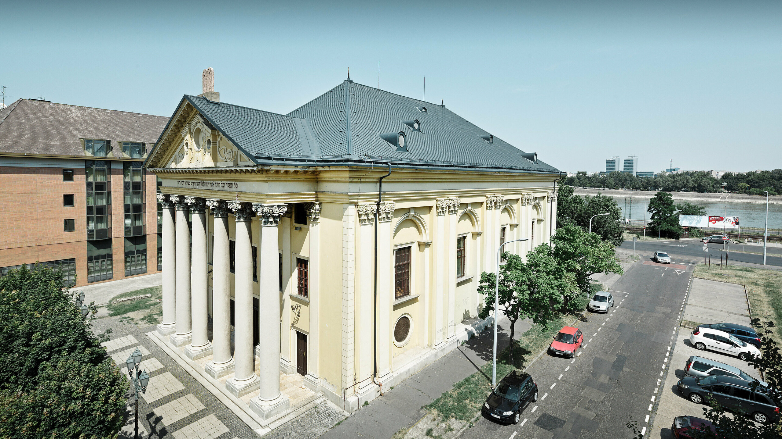 Historisches Gebäude mit klassizistischer Architektur und heller Fassade in Cremegelb. Das Bauwerk besitzt ein dunkelgraues Walmdach aus Aluminiumrauten und wird von einer Reihe großer Säulen an der Vorderseite geprägt. Es steht an einer Straße mit parkenden Autos, nahe eines Flusses, mit modernen Gebäuden im Hintergrund.