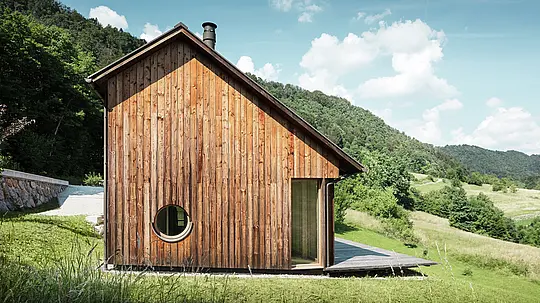 Minimalistisches Holzhaus mit Rundfenster und robustem PREFALZ-Aluminiumdach in der Farbe P.10 Braun, eingebettet in die sanfte Hügellandschaft von Vinje. Die wartungsfreie PREFA-Dachdeckung bietet optimalen Schutz vor Wind und Wetter und harmoniert farblich perfekt mit der natürlichen Holzfassade. Sie unterstreicht die klare, zeitlose Architektur des Gebäudes. Die großzügige Terrasse eröffnet einen herrlichen Blick auf die Hügellandschaft Sloweniens. Die Perspektive zeigt die Fassade des Einfamilienhauses mit asymmetrischem Satteldach und unterschiedlich großen Dachflächen.