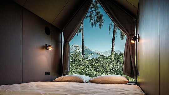 Blick aus dem Bett eines minimalistischen Tiny Houses im Secret Koi Garden am Natterer See: Durch die große, dreieckige Glasfront öffnet sich der Raum zur Natur mit Bäumen und Blick auf die Tiroler Berglandschaft. Das reduzierte Interior lenkt den Fokus nach außen und unterstreicht das Konzept der Entschleunigung. Geschützt wird die Architektur von langlebigen PREFA Aluminiumrauten, die Design, Wetterbeständigkeit und Nachhaltigkeit vereinen.