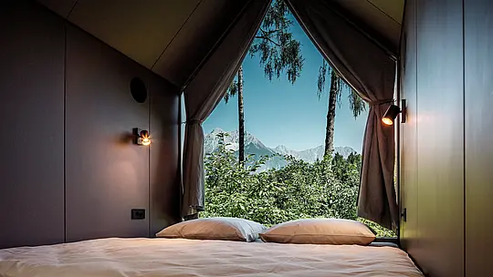 Blick aus dem Bett eines minimalistischen Tiny Houses im Secret Koi Garden am Natterer See: Durch die große, dreieckige Glasfront öffnet sich der Raum zur Natur mit Bäumen und Blick auf die Tiroler Berglandschaft. Das reduzierte Interior lenkt den Fokus nach außen und unterstreicht das Konzept der Entschleunigung. Geschützt wird die Architektur von langlebigen PREFA Aluminiumrauten, die Design, Wetterbeständigkeit und Nachhaltigkeit vereinen.