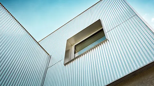 Die markante PREFA Strangpressprofilfassade in der Ausführung "Welle” setzt das Envirocenter Nitra eindrucksvoll in Szene. Aus der Froschperspektive entfalten die vertikalen Aluminiumprofile in Naturblank ihre dynamische Wellenstruktur und reflektieren das Sonnenlicht. Ein präzise eingefasstes Fenster durchbricht die silbern schimmernde Gebäudehülle. Die langlebigen und witterungsbeständigen Aluminiumprofile vereinen moderne Ästhetik mit hoher Funktionalität.