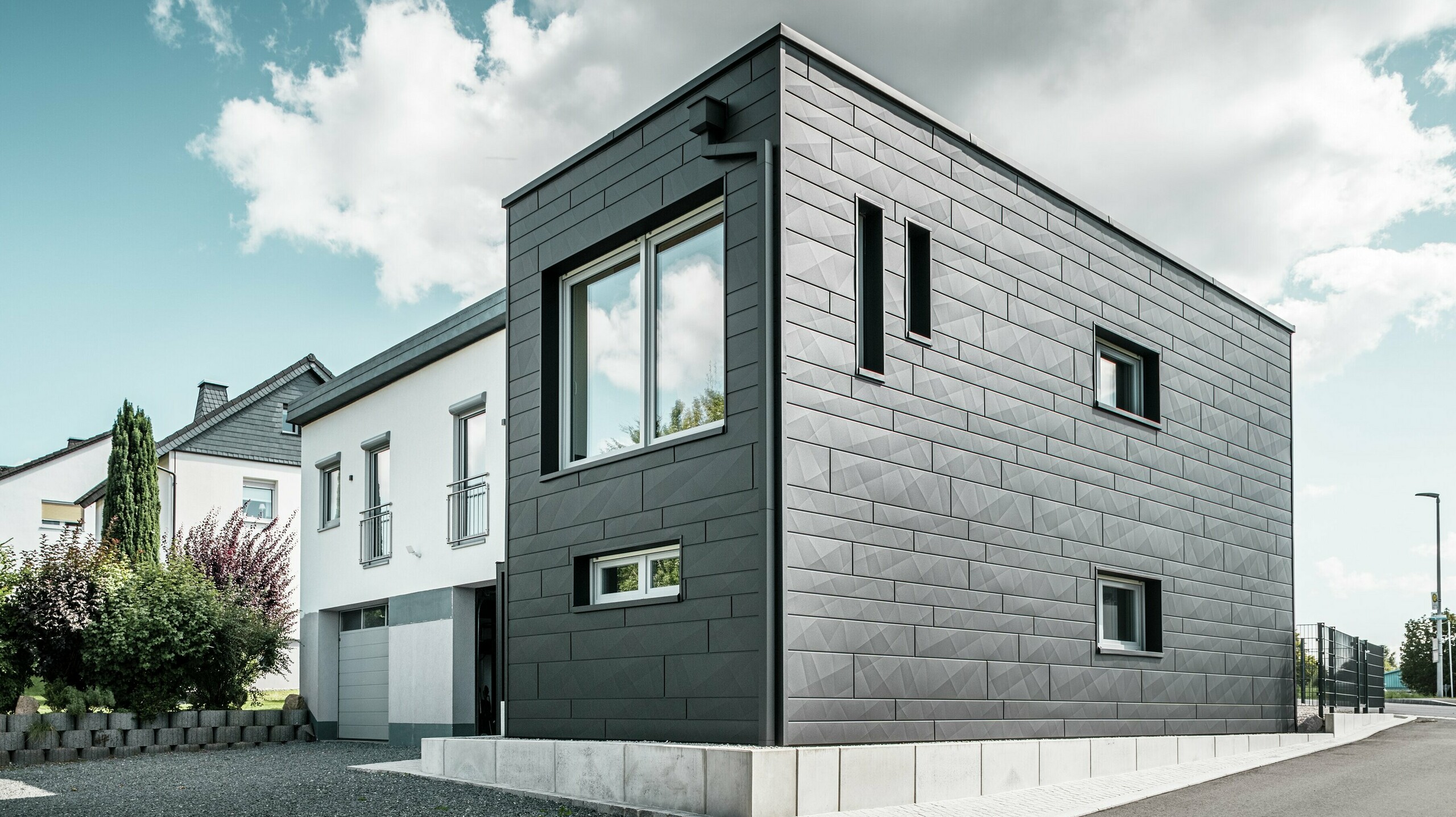 Die stilvolle und moderne Fassade des sanierten Einfamilienhauses in Teuschnitz zeigt die klar strukturierten PREFA Siding.X Paneele in P.10 Schwarz. Die geometrische Anordnung der hochwertigen Blechpaneele verleiht dem Gebäude eine markante, zeitlose Ästhetik. Die Fenster sind von schwarzen Aluminium Verbundplatten umrahmt, was das Design harmonisch abrundet. Die integrierte PREFA Dachentwässerung mit dem Quadratrohr sorgt für eine funktionale und ästhetische Lösung.