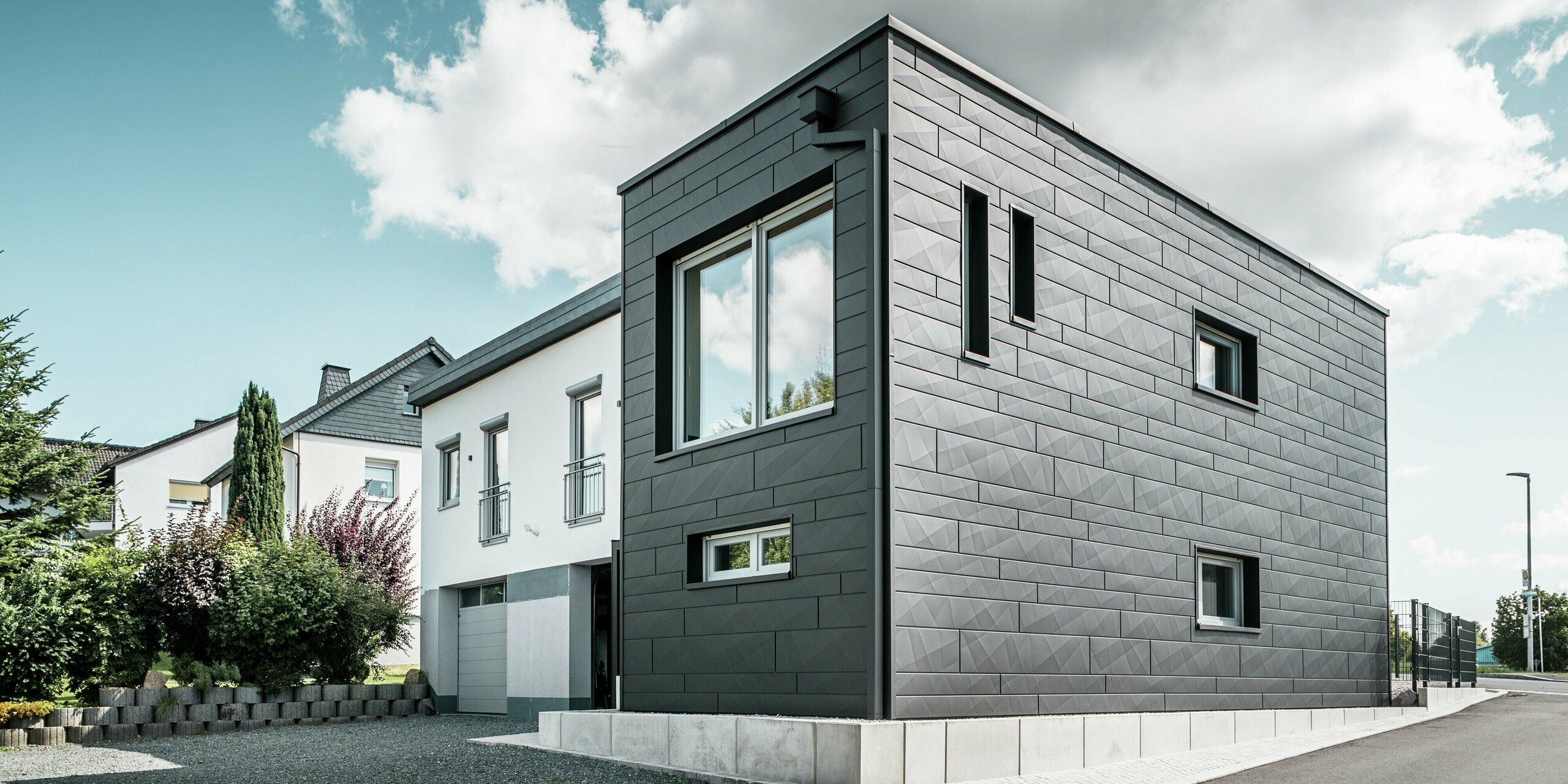 Die stilvolle und moderne Fassade des sanierten Einfamilienhauses in Teuschnitz zeigt die klar strukturierten PREFA Siding.X Paneele in P.10 Schwarz. Die geometrische Anordnung der hochwertigen Blechpaneele verleiht dem Gebäude eine markante, zeitlose Ästhetik. Die Fenster sind von schwarzen Aluminium Verbundplatten umrahmt, was das Design harmonisch abrundet. Die integrierte PREFA Dachentwässerung mit dem Quadratrohr sorgt für eine funktionale und ästhetische Lösung.