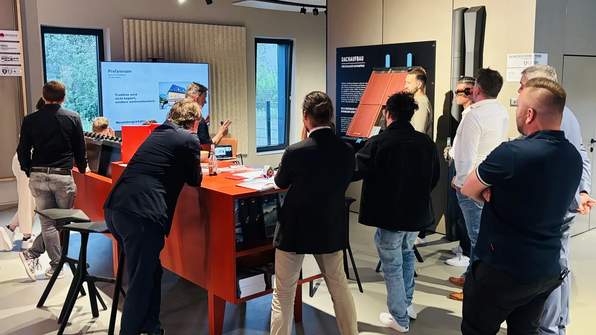 Architektur- und Planungspartner:innen versammeln sich im PREFA Showroom St. Veit an der Glan um eine Präsentationsstation mit digitalen Inhalten und Mustermaterialien. Auf einem Bildschirm werden Referenzen moderner Dach- und Fassadenprojekte gezeigt, während ein PREFA Aluminium-Dachsystem anschaulich erläutert wird. Die Teilnehmer:innen verfolgen die Inhalte im Barcamp-Format und tauschen sich direkt vor Ort aus. Die Szene zeigt praxisnahe Wissensvermittlung, innovative PREFA Aluminiumlösungen und interaktive Einblicke in Gestaltungsmöglichkeiten für zeitgemäße Dach- und Fassadenplanung.