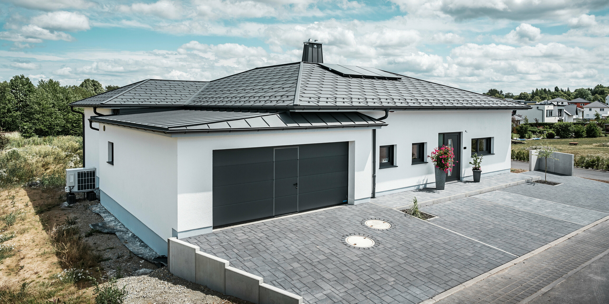 Modernes Wohndesign vereint mit Funktionalität in Ilmenau, Thüringen: Dieser ansprechende Bungalow präsentiert sich mit einem dunkelgrauen PREFA Aluminiumdach, gekennzeichnet durch die stilvollen Dachplatten R.16. Die Garage fügt sich mit PREFALZ in gleicher Farbgebung nahtlos in die Gesamtarchitektur ein. Der Schornstein mit dem PREFA Stier-Symbol ragt markant empor, während das Dachentwässerungssystem mit Ablaufrohr und Hängerinne eine zuverlässige und ästhetische Lösung zur Wasserableitung darstellt.