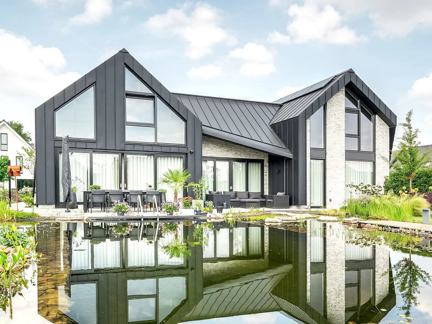 Modernes Einfamilienhaus mit charakteristischen, spitzen Giebeldächern, bedeckt mit schwarzen Prefalz-Dachbahnen, reflektiert sich in einem ruhigen Gartenteich. Die Fassade kombiniert dunkle Wandelemente mit hellen Ziegelbereichen und großen Fenstern. Eine Terrasse mit Gartenmöbeln lädt zum Verweilen ein und ist harmonisch in die üppige, blühende Gartenlandschaft integriert.