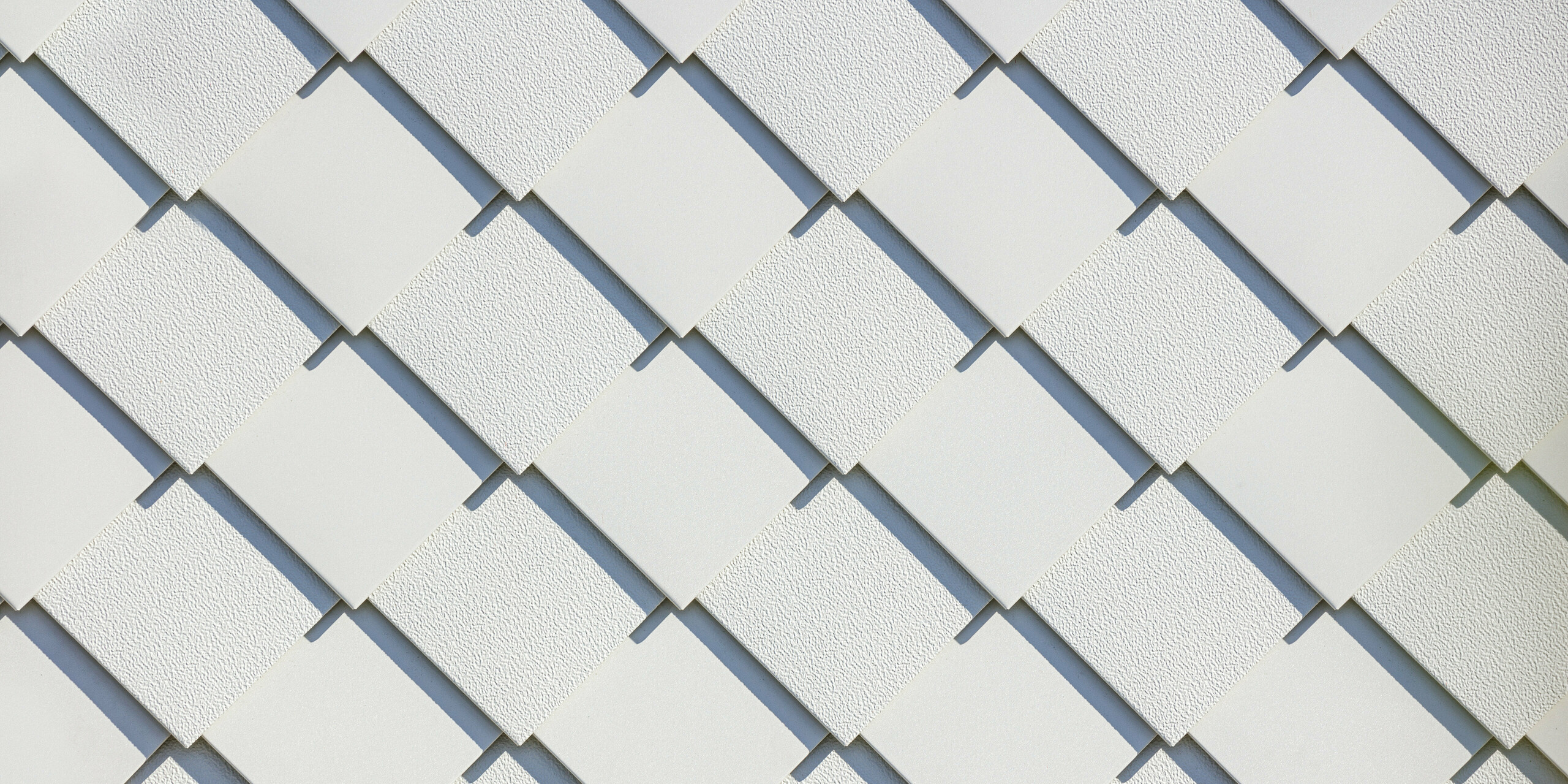 Nahaufnahme der PREFA Wandrauten 20x20 in P.10 Prefaweiß, die als Fassadensystem an einem Neubau-Mehrfamilienhaus in der Schweiz angebracht sind. Die vielseitige Oberflächengestaltung der Aluminiumrauten wurde hierbei als Designelement genutzt. Die Linien der Rauten wurden abwechselnd mit glatter und mit Stucco-Oberfläche verlegt. Die wechselnde Oberflächenstruktur betont die präzise Verarbeitung zusätzlich. Die PREFA Dach- und Wandrauten sind als langlebige und wartungsarme Fassadenlösung bekannt und bieten durch ihre gleichmäßige Anordnung eine elegante Ästhetik.