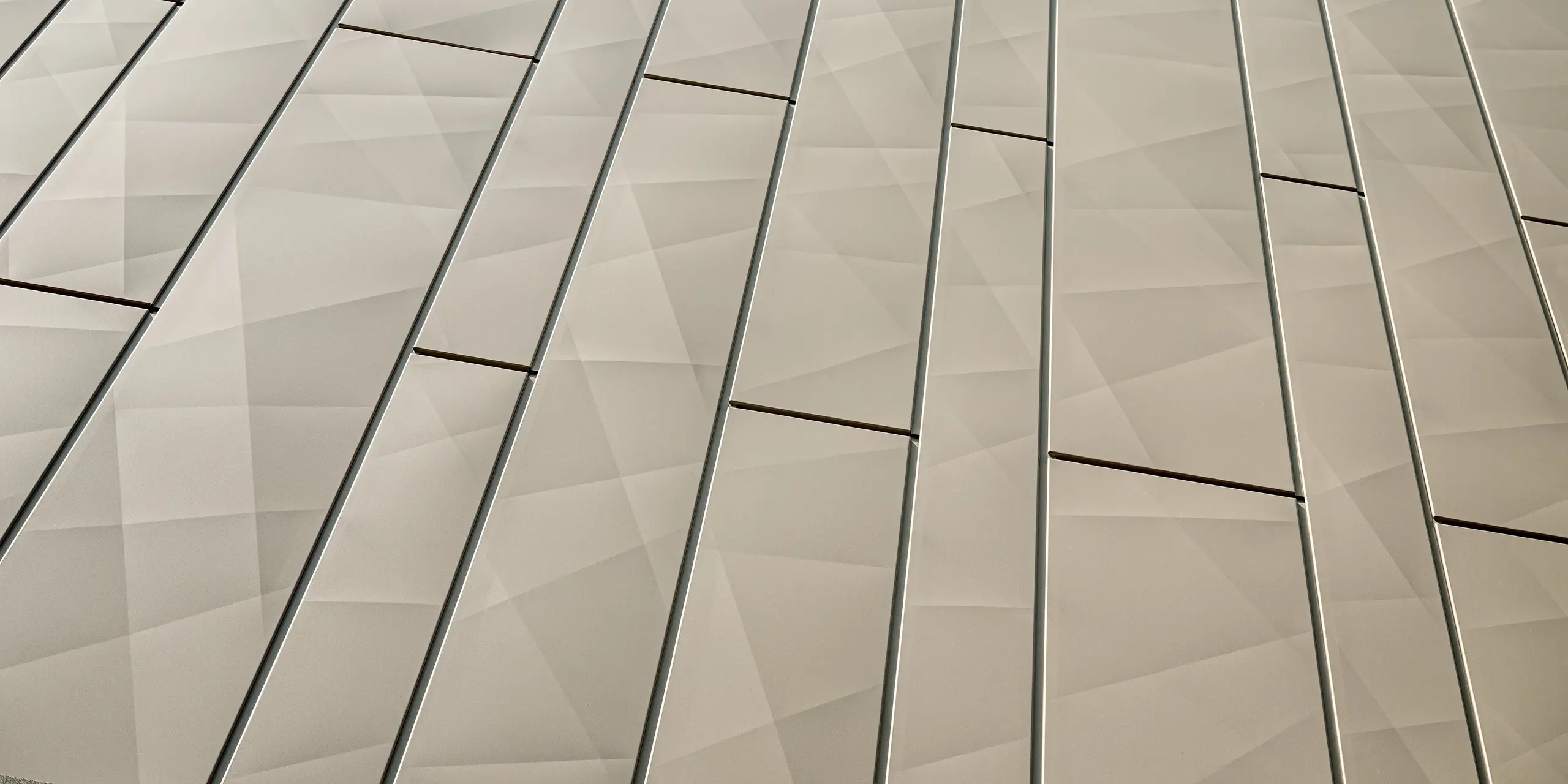 Das Bild zeigt einen Nahaufnahme der Siding-X-Paneele der Aluminiumfassade des Einkaufszentrums in Rohrbach. Die in einem eleganten Bronzeton gehaltenen Fassadenelemente zeichnen sich durch eine geometrische Struktur aus, die durch präzise Linien und flache Falten eine dynamische, dreidimensionale Textur erzeugt. Die nahtlosen Übergänge und die feine Unterteilung der Paneele vermitteln eine moderne, elegante Optik. Diese Fassadenelemente sind nicht nur ästhetisch ansprechend, sondern auch äußerst widerstandsfähig gegen äußere Einflüsse, wodurch sie besonders pflegeleicht und langlebig sind.