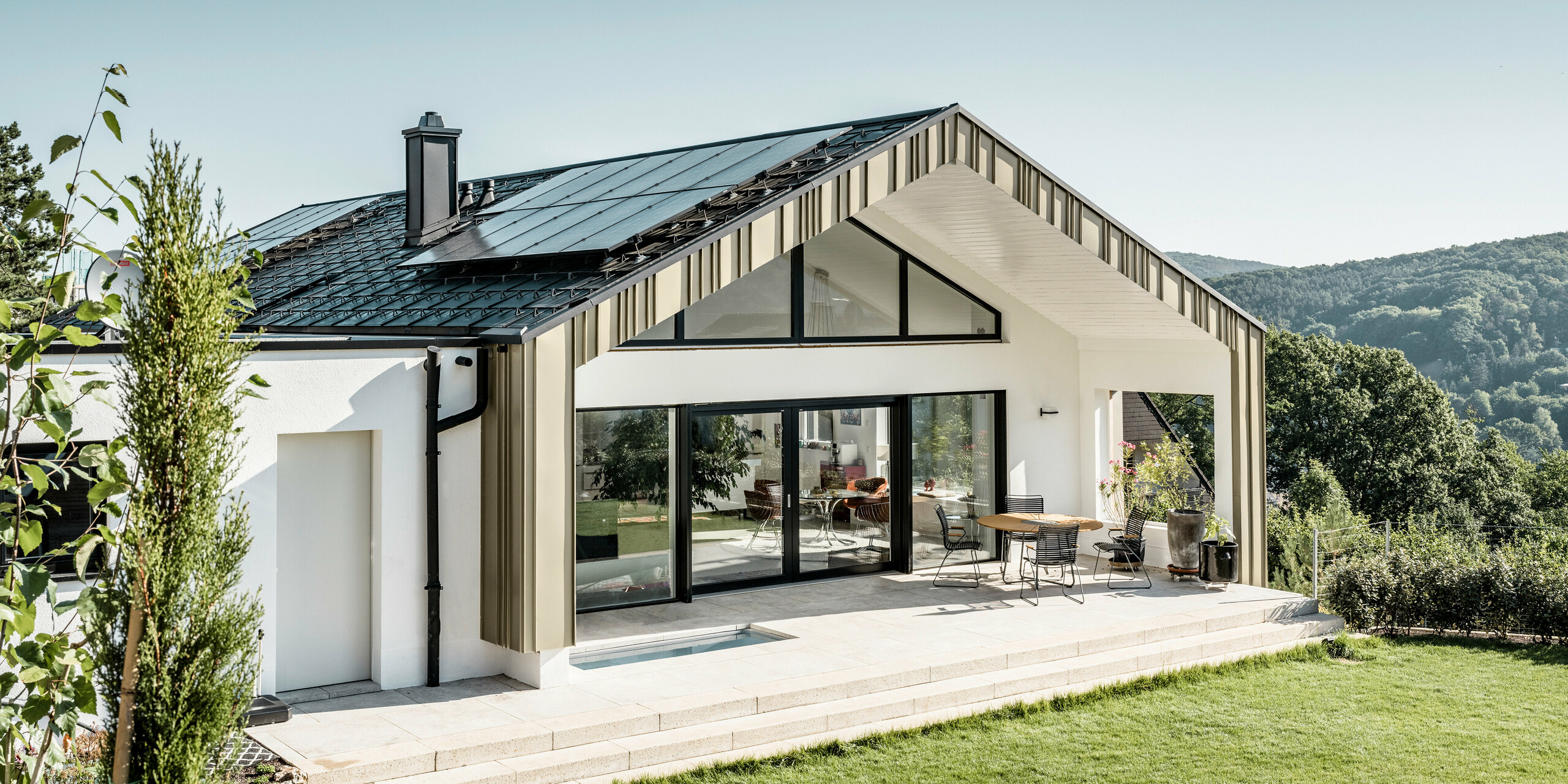 Modernes Einfamilienhaus mit einem langlebigen PREFA Aluminiumdach in Anthrazit, das mit der hochwertigen Dachplatte R.16 ausgestattet ist. Die elegante PREFA Fassade in Falzonal Lichtbronze ist perfekt integriert in eine idyllische, grüne Landschaft. Die umweltfreundliche und energieeffiziente Bauweise wird durch die große Glasfront und die harmonische Integration in den natürlichen Hintergrund unterstrichen, was die Ästhetik und Funktionalität von PREFA Dach- und Fassadensystemen betont. 