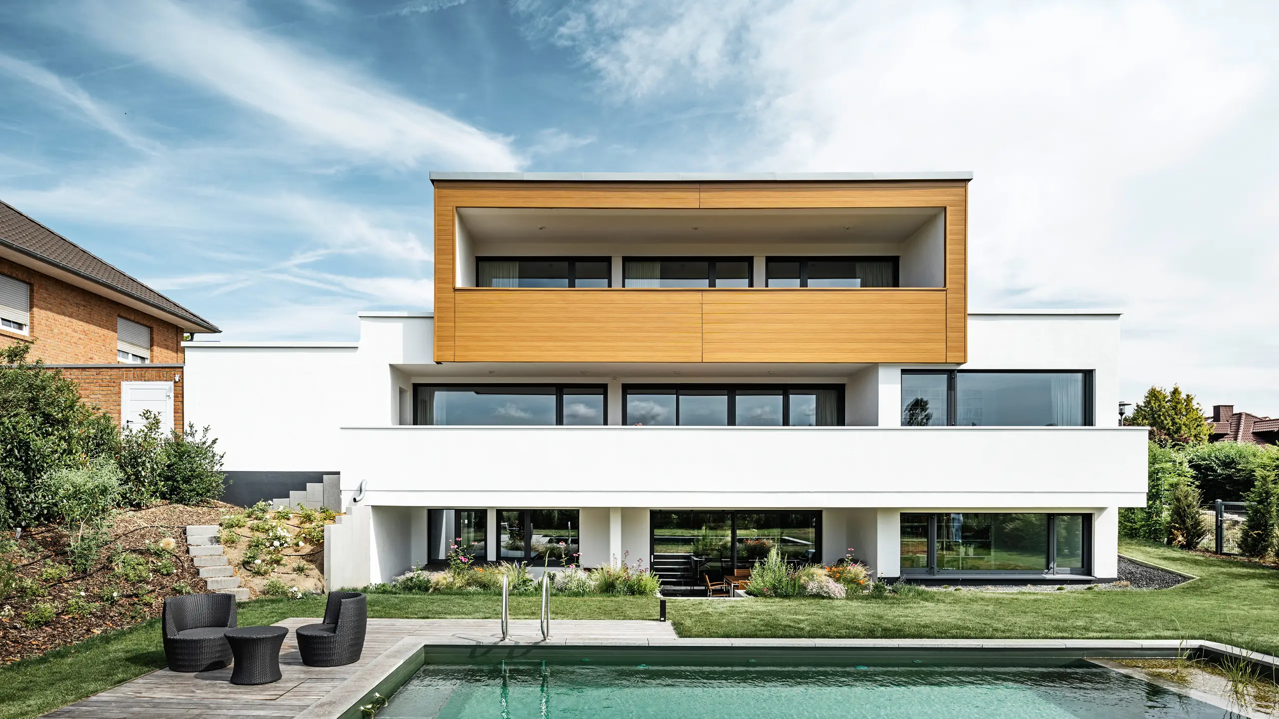 Die Rückseite eines Einfamilienhauses in Usingen, Deutschland. Am unteren Bildrand ist der gepflegte Pool und Garten inkl. Outdoor-Sitzmöbel zu sehen. Das oberste Geschoss des Gebäudes wurde mit PREFA Sidings in Eiche natur umhüllt. Der Gebäudesockel wurde mit einer weißen Putzfassade ausgestattet.
