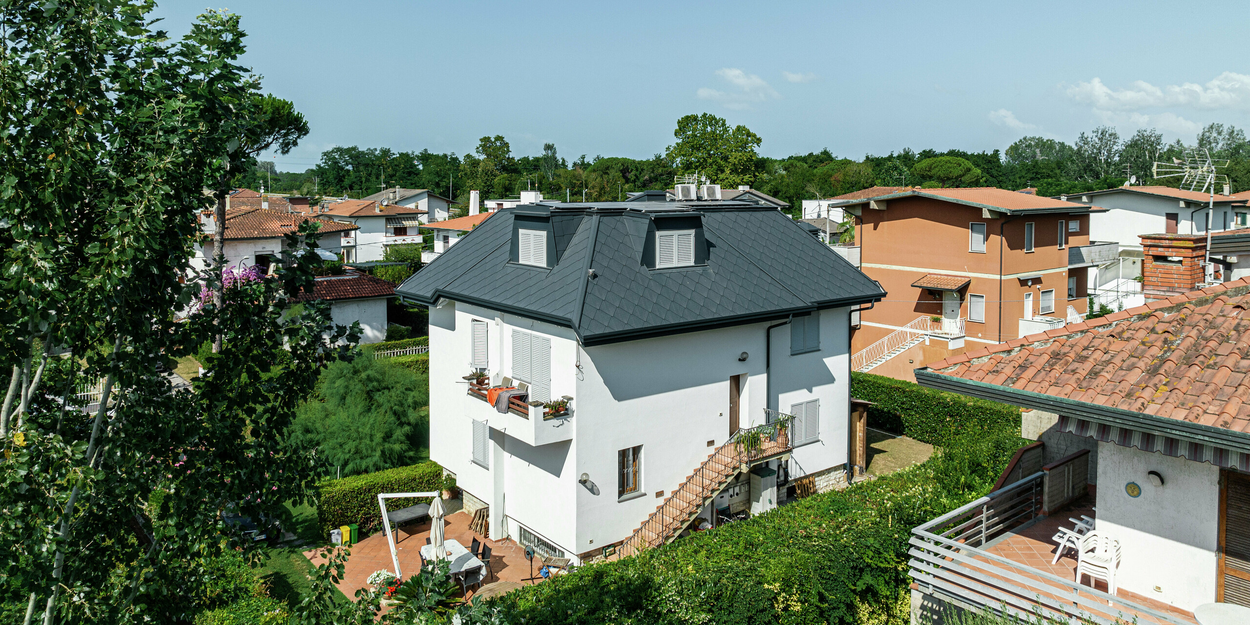 Stilvolles Einfamilienhaus in Montignoso aus einer erhöhten Perspektive. Die PREFA Dachraute 44 × 44 in P.10 Steingrau verleiht dem Dach eine elegante Optik und überzeugt durch Langlebigkeit und extreme Witterungsbeständigkeit. Die dunkle Dacheindeckung bildet einen schönen Kontrast zur weißen Fassade. Die Gauben sind harmonisch in die Dachkonstruktion integriert.