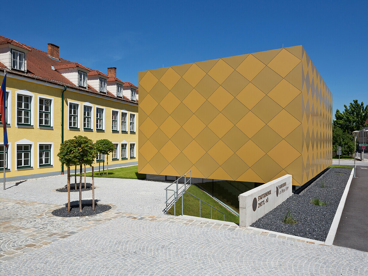 Blick aus der Distanz auf den Eingangsbereich des Musikvereins in Zwettl. Das quaderförmige Gebäude steht vor dem Stadtamt in Zwettl und sticht durch die markante Hülle aus PREFA Aluminium Verbundplatten hervor.