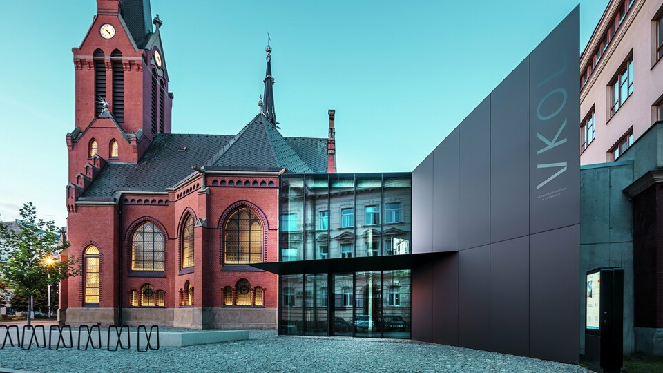 Das Bild zeigt das imposante Zusammenspiel zwischen der historischen Roten Kirche und dem modernen Neubau in Olmütz. Die Kirche mit ihren charakteristischen gotischen Fenstern und dem markanten Turm bildet das historische Rückgrat des Ensembles, während der moderne Anbau mit seiner eleganten, schwarzen PREFABOND Aluminium-Verbundplatten-Fassade eine zeitgemäße, architektonische Antwort darstellt. Große Glasflächen lassen den Blick in das Innere des Neubaus zu, wo die Umgebung durch Reflexionen in den Fenstern eine interessante Wechselwirkung zwischen Alt und Neu widerspiegelt. Die präzise Verarbeitung der PREFABOND-Platten betont die moderne Ästhetik, während die harmonische Integration in das historische Umfeld sowohl funktional als auch visuell beeindruckt.