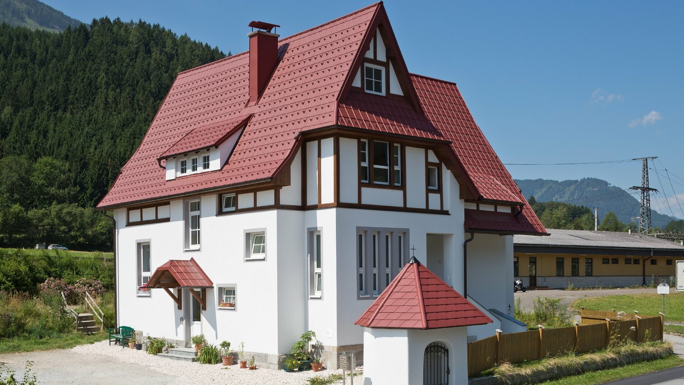Verwinkeltes Steildach mit PREFA Dachplatte in P.10 Oxydrot mit vielen Details wie Gauben, Türmchen und Kamineinfassungen. Auch das Vordach und das Marterl vor dem Haus sind mit PREFA Blechdach eingedeckt.