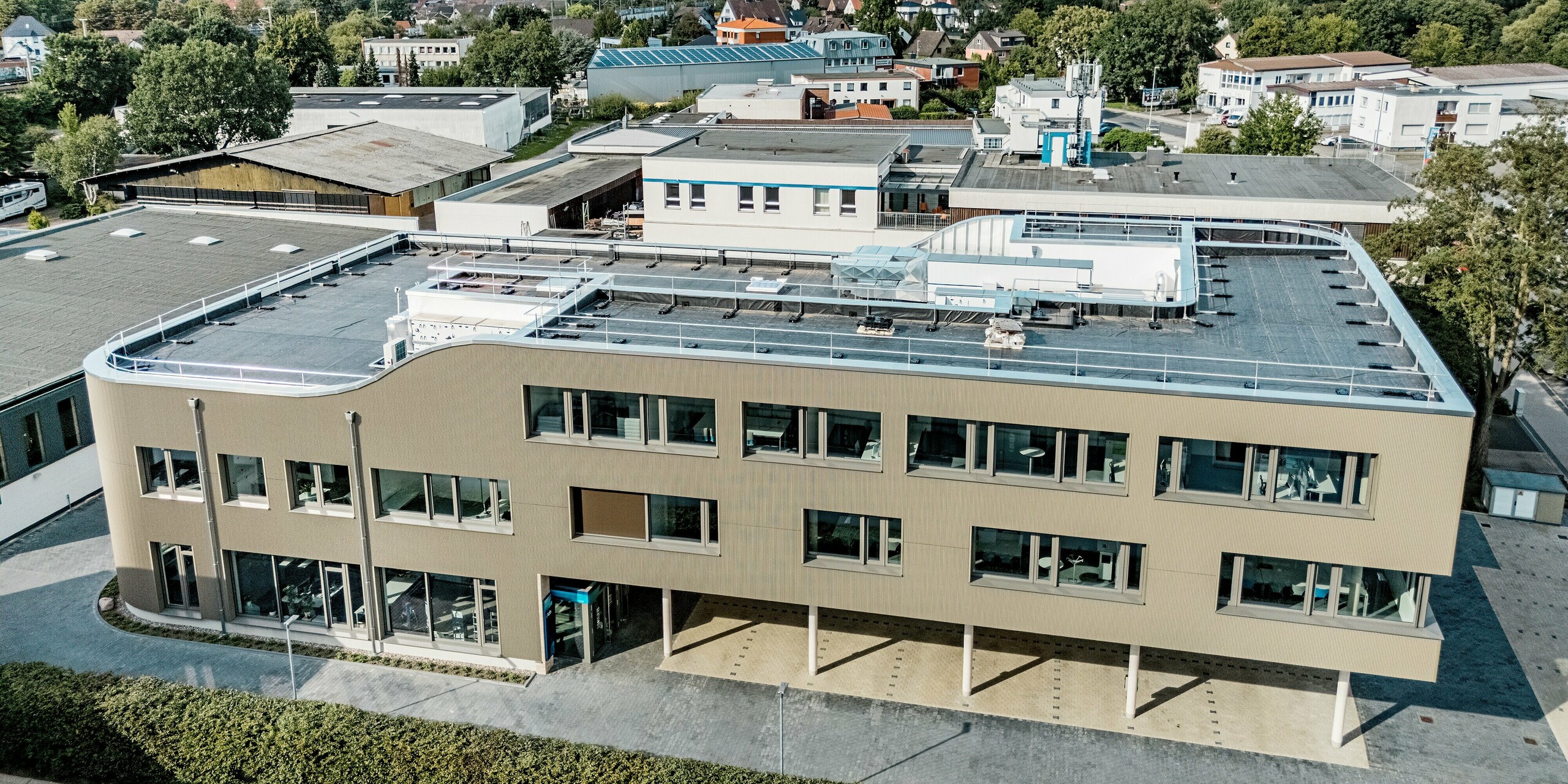 Das Bild zeigt das Festo-Gebäude in Langenhagen aus der Vogelperspektive. Die markante Fassade aus PREFA Strangpressprofilen in Zackenform in Bronze verleiht dem Gebäude eine moderne und dynamische Ausstrahlung. Besonders auffällig ist die geschwungene Architektur der oberen Fassadenkante, die dem Gebäude eine elegante und zeitgemäße Form verleiht. Die großen Fensterflächen und die klar strukturierte Außenwand bieten eine harmonische Kombination aus Funktionalität und ästhetischem Design. Durch die Verwendung von PREFA Produkten wird eine lange Lebensdauer sowie eine ausgezeichnete Widerstandsfähigkeit gegen äußere Einflüsse garantiert. Die präzise Verarbeitung und hohe Qualität der Materialien machen das Gebäude zu einem herausragenden Beispiel für moderne Architektur.