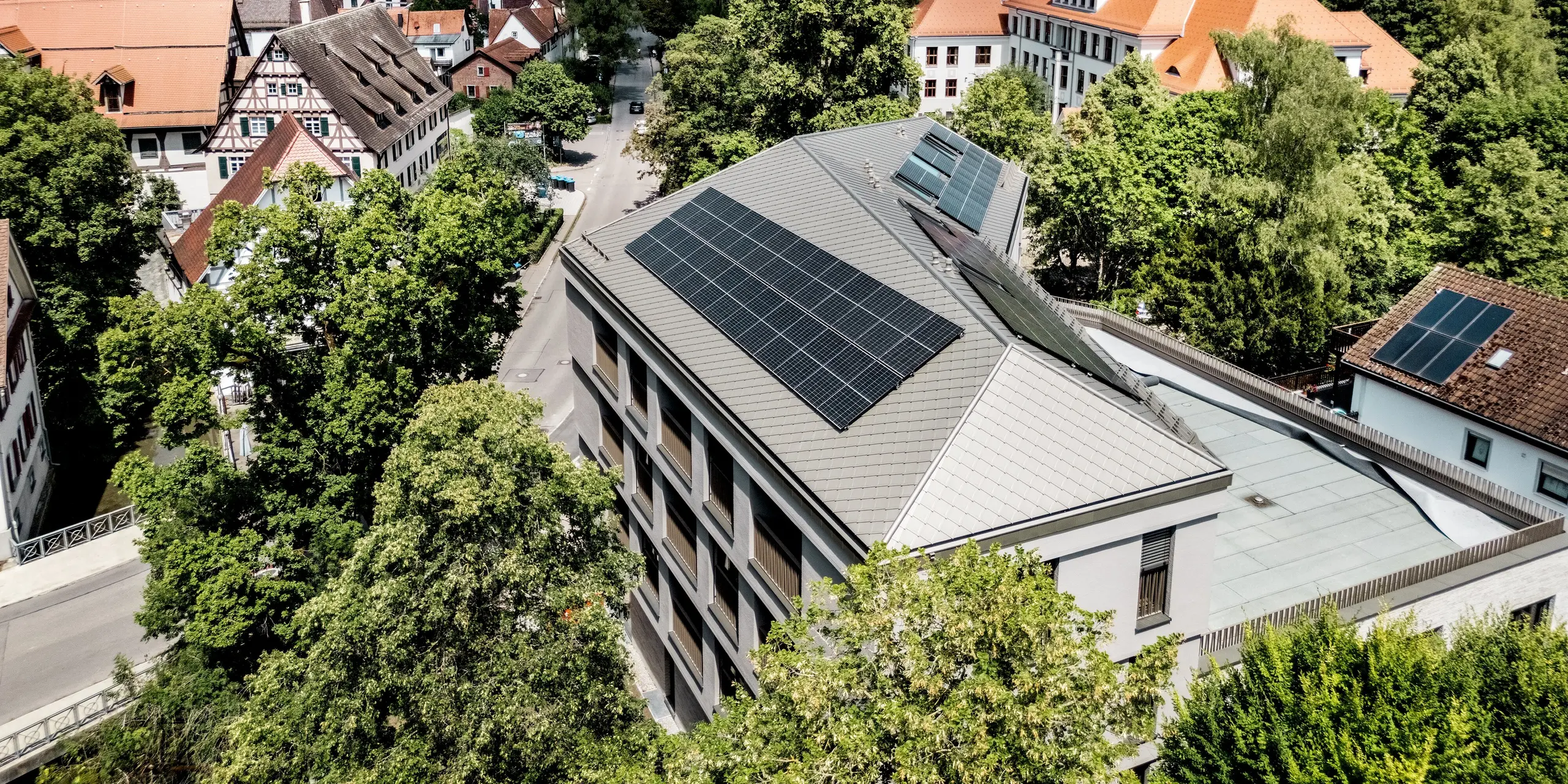 Beeindruckende Luftaufnahme der Sparkasse Blaubeuren inmitten der historischen Altstadt mit malerischen Fachwerkhäusern und grünen Hügeln. Das hochwertige Blechdach mit PREFA Dachrauten 44 × 44 in Bronze und großflächigen Photovoltaikanlagen setzt einen modernen Akzent. Die leichten, langlebigen und witterungsbeständigen Aluminiumrauten vereinen elegante Architektur mit hoher Funktionalität.
