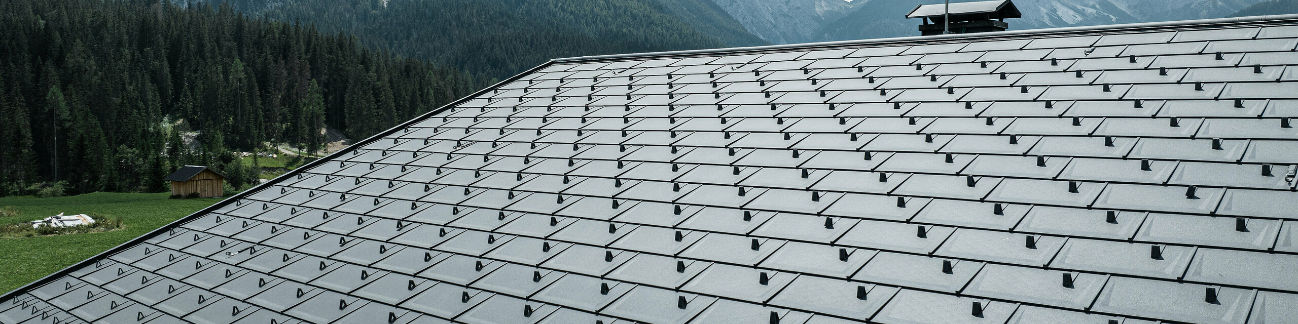 Casa Hoffe in Sappada, Italien, geprägt durch die PREFA Dachplatten R.16 in P.10 Schwarz. Das Dachsystem fügt sich hervorragend in die natürliche Umgebung der Dolomiten ein. Die präzise verlegten Dachplatten bieten eine effiziente und dauerhafte Bedeckung, die das traditionelle Erscheinungsbild des Hauses mit modernster Technik und Materialbeständigkeit vereint. Der Anblick des robusten Aluminiumdachs vor dem Hintergrund der beeindruckenden Bergkulisse symbolisiert die Verschmelzung von Natur und fortschrittlicher Bautechnologie.