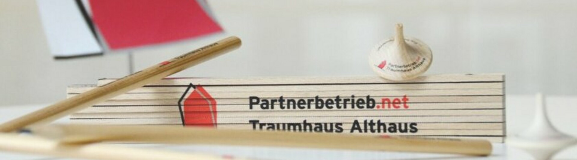Zollstock mit Aufdruck „Partnerbetrieb.net Traumhaus Althaus“ auf einem Tisch, umgeben von Holzstäben und einem Kreisel, im Hintergrund ein Schild mit Haus-Logo.