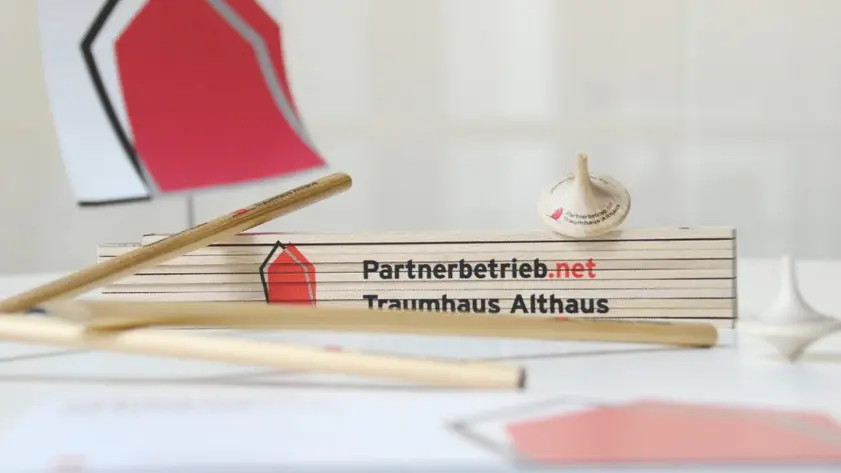 Zollstock mit Aufdruck „Partnerbetrieb.net Traumhaus Althaus“ auf einem Tisch, umgeben von Holzstäben und einem Kreisel, im Hintergrund ein Schild mit Haus-Logo.
