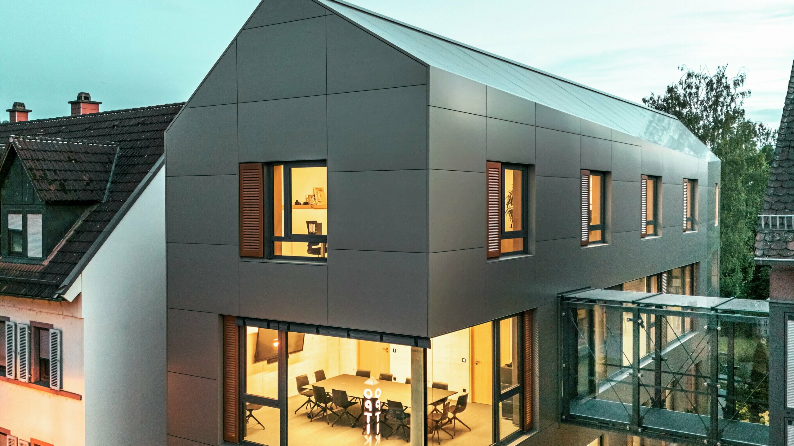 Das Bild zeigt das Rathaus in Alzenau bei Dämmerung, mit einer modernen Fassade aus Aluminium-Verbundplatten von PREFA in einem eleganten Bronze-Ton. Der gläserne Verbindungsgang, der das Gebäude mit einem angrenzenden Teil verbindet, hebt sich visuell ab. Die großen Fenster gewähren Einblicke in den Innenbereich, der durch warmes Licht erleuchtet wird. Die klare geometrische Form der Fassade und die umgebende Grünanlage verstärken den modernen, zeitgemäßen Look des Gebäudes.
