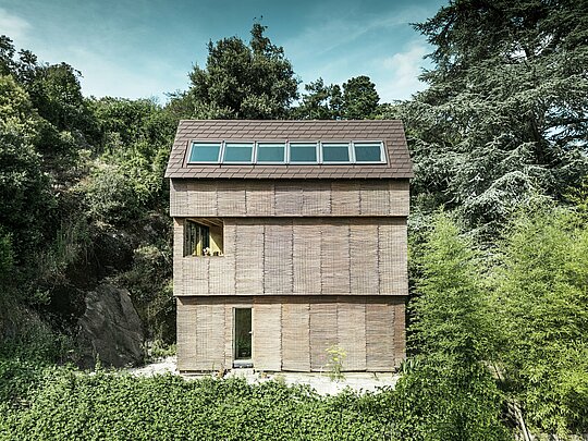 Moderne Architektur in Frankreich – Haus mit PREFA Dachschindeln in P.10 Nussbraun und Weidenfassade, steil gelegen auf Hügel mit großzügigen Dachfenstern.