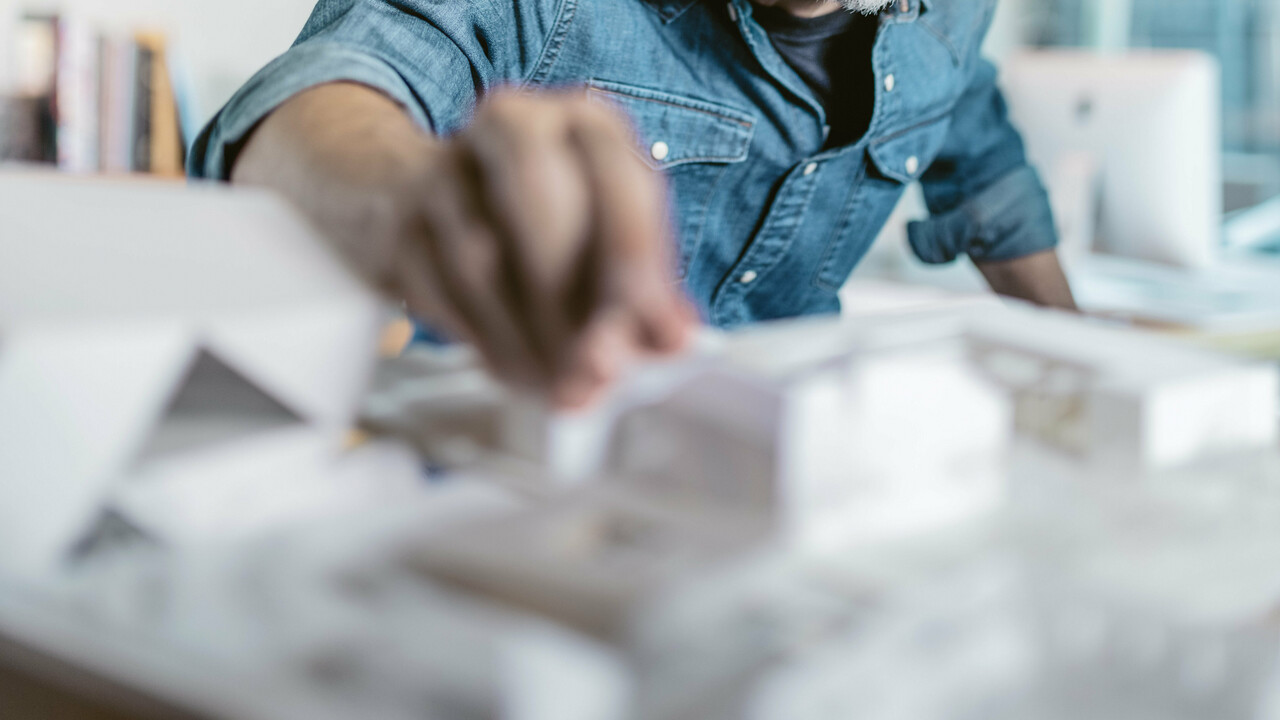 Architekt bei der Arbeit mit einem Architekturmodell, umgeben von Skizzen und Entwürfen, während er die Details eines Projekts prüft. Das Bild vermittelt Kreativität und Präzision, passend zur Bewerbung der hochwertigen Aluminiumprodukte und Farbmuster von PREFA, die für Architekten maßgeschneiderte Lösungen bieten.