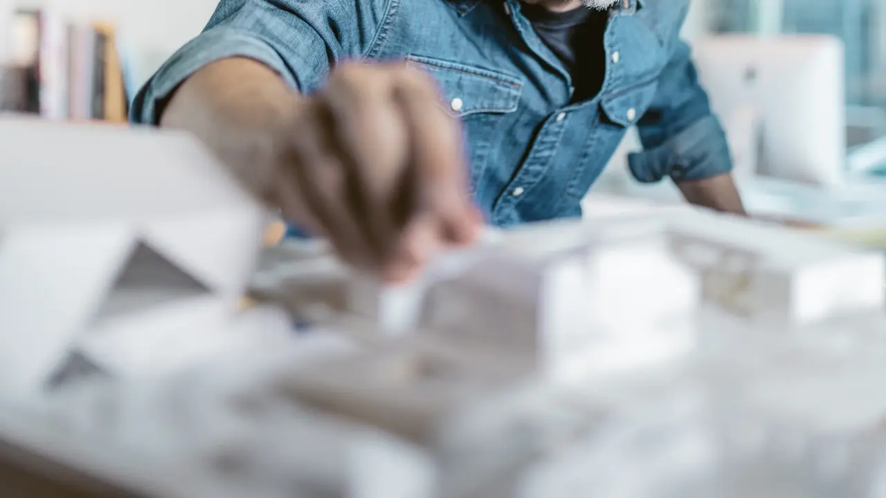 Architekt bei der Arbeit mit einem Architekturmodell, umgeben von Skizzen und Entwürfen, während er die Details eines Projekts prüft. Das Bild vermittelt Kreativität und Präzision, passend zur Bewerbung der hochwertigen Aluminiumprodukte und Farbmuster von PREFA, die für Architekten maßgeschneiderte Lösungen bieten.