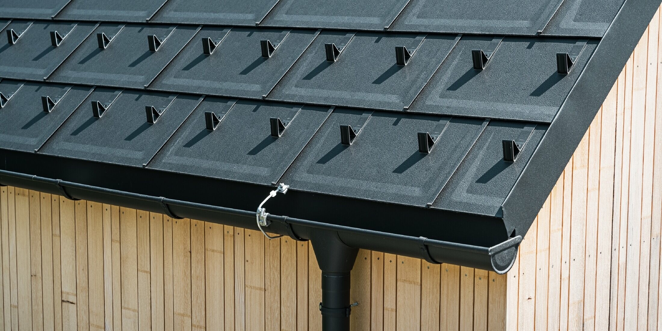 Nahaufnahme der PREFA Dachplatte R.16 mit Schneestoppern, Dachrinne und Ablaufrohr in Schwarz P.10 inkl. Blitzschutz und einer senkrechten Holzfassade
