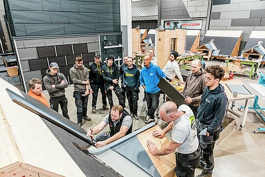 Eine Gruppe von Fachleuten, die an einer Schulung in der PREFA Academy teilnehmen. Ein PREFA Lehrverleger arbeitet an einem Dachmodell, während die anderen aufmerksam zuschauen. Im Hintergrund sind verschiedene Dachmodelle und Arbeitsplätze sichtbar.