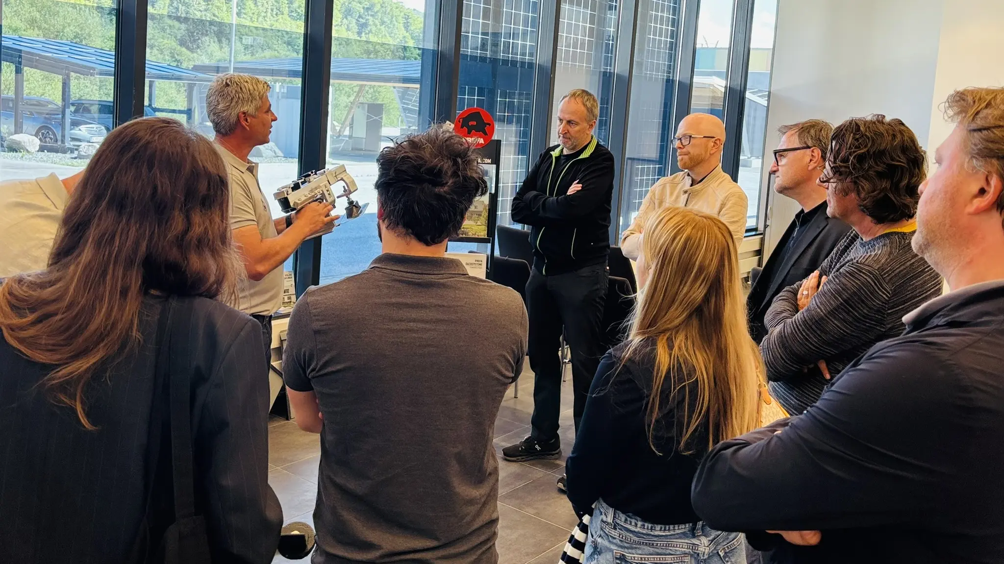 Teilnehmende des PREFA Barcamp-Abends im Showroom St. Veit an der Glan verfolgen aufmerksam eine technische Präsentation zu Aluminium-Dach- und Fassadenlösungen. Eine Person demonstriert ein Werkzeug oder Bauteil, während eine Gruppe von Architektur- und Planungspartner:innen im Halbkreis zuhört. Große Fensterflächen geben den Blick auf außenliegende Solarmodule frei, die den Bezug zu nachhaltigen PREFA Systemlösungen unterstreichen. Die Szene zeigt intensiven fachlichen Austausch, praxisnahe Einblicke und die direkte Vermittlung von Know-how rund um moderne Dach- und Fassadenprojekte.