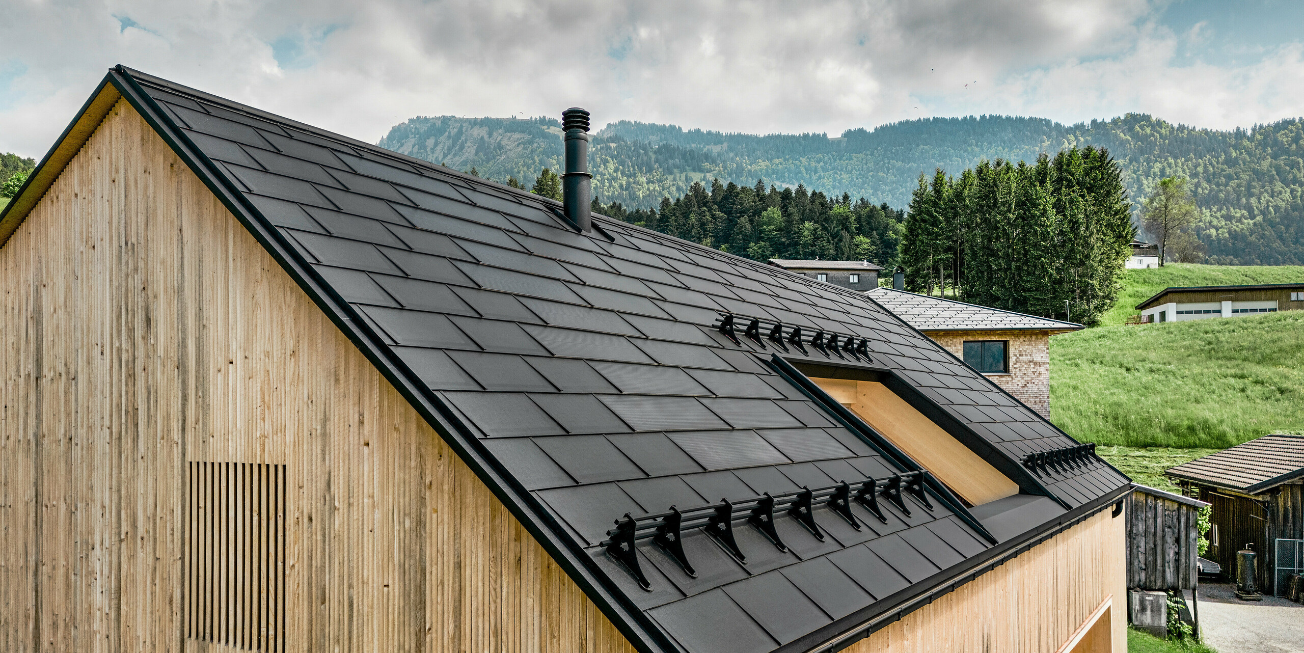 Detailaufnahme eines ökologisch konzipierten Hauses in Egg, ausgestattet mit effizienten PREFA Solardachplatten, die sich zwischen den hochwertigen schwarzen PREFA Dachplatten R.16 hervorheben. Das natürliche Holz der Fassade bildet einen warmen Kontrast zum technologischen Flair des schwarzen Aluminiumdaches, das Energieeffizienz mit stilvoller Ästhetik verbindet. Das Dach, gekrönt mit einem schlanken Schornstein, spiegelt eine nachhaltige Bauweise wider, die in der ländlichen Umgebung mit sanften Hügeln und üppigem Grün verwurzelt ist.