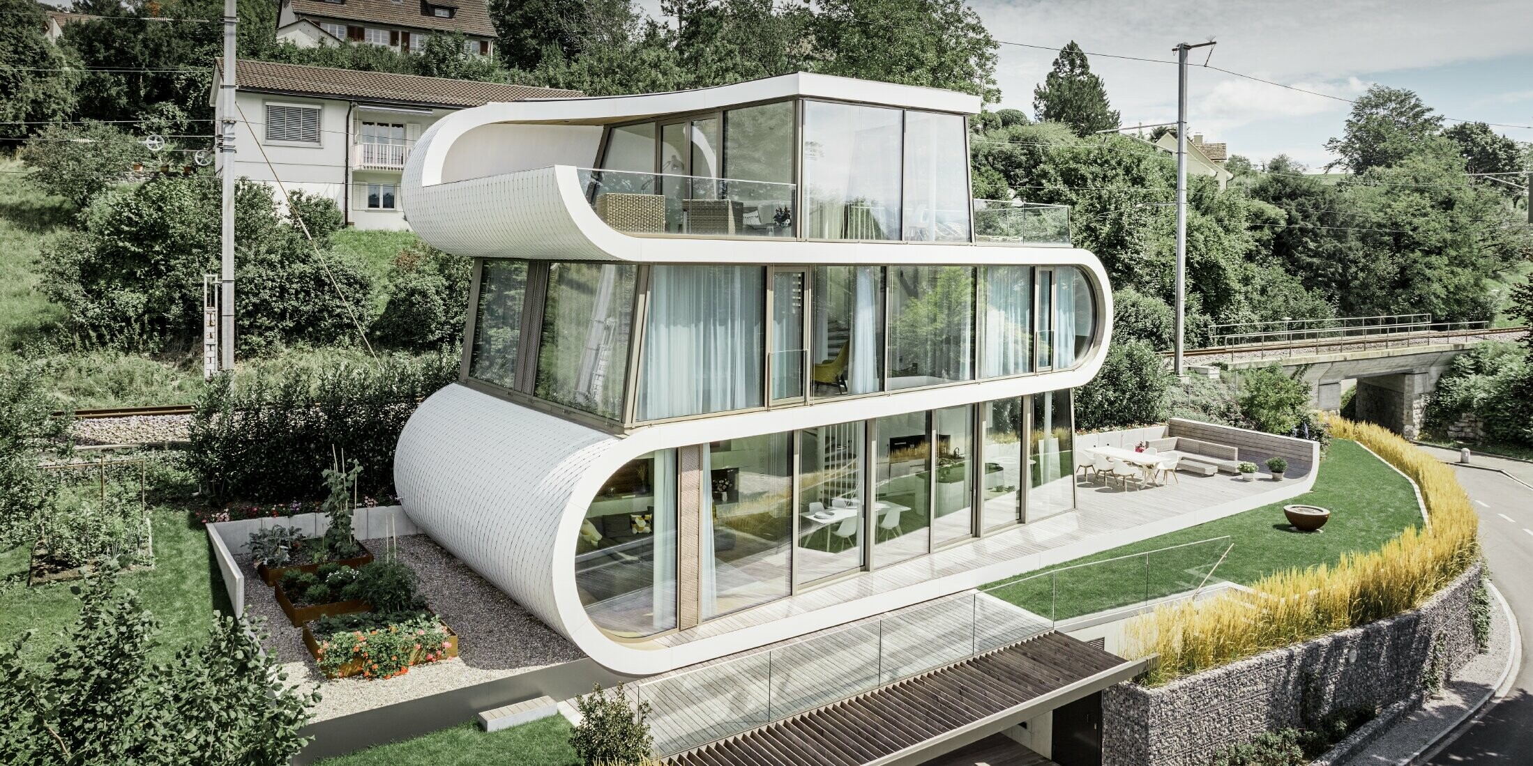 Sehr modernes Einfamilienhaus entworfen durch Architekt Camenzind aus Zürich; Ein geschwungenes Band verbindet die einzelnen Ebenen miteinander. Die Rundungen wurden mit der PREFA Dachraute klein in Reinweiß verkleidet. Das Haus hat viele und große Glasflächen, wodurch das ganze Haus sehr offen wirkt.