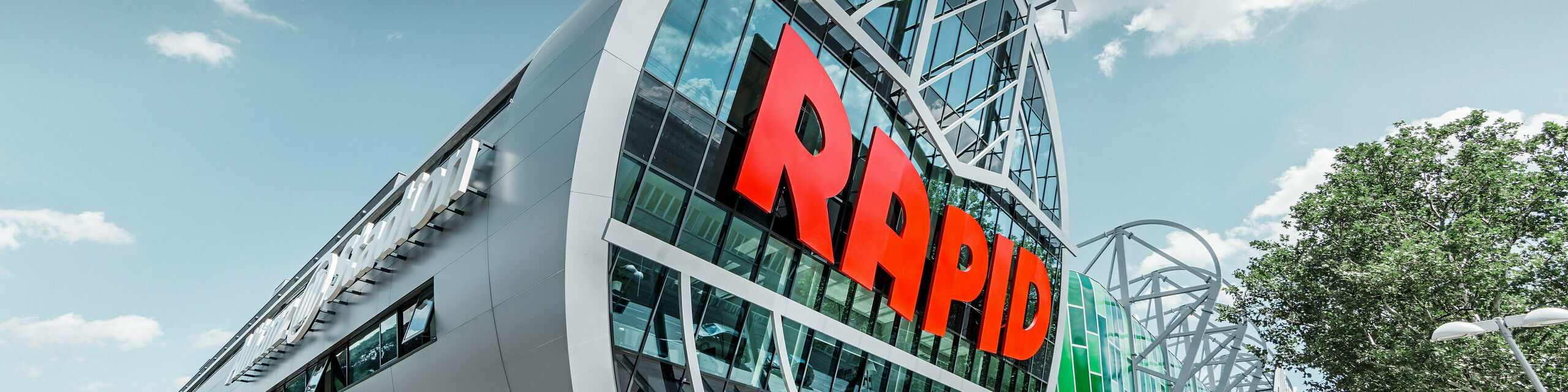 Ansicht des Allianz Stadions von SK Rapid Wien mit auffälliger PREFA Fassade in Form der Aluminium Verbundplatte in Silbermetallic. Die Gebäudehülle strahlt Dynamik und Moderne aus. Der Eingangsbereich, gekrönt mit dem leuchtend roten RAPID-Schriftzug, lädt ein in das Herz des Fußballs, eingefasst von einer futuristischen und zugleich eleganten Fassadenstruktur, die Sportarchitektur auf ein neues Niveau hebt.