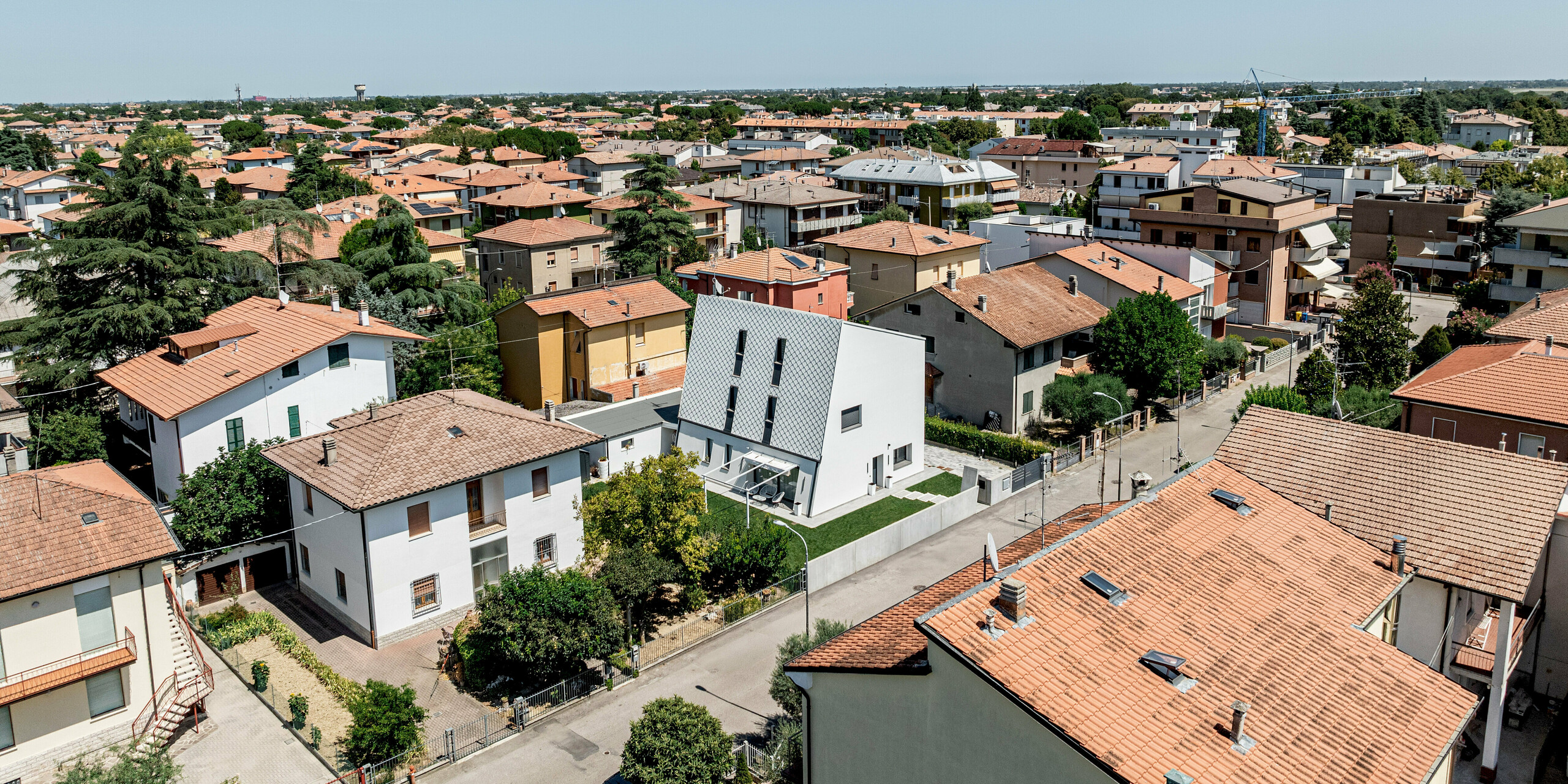 Weitläufige Luftaufnahme eines Wohnviertels in Forlì, Italien, mit einem modernen Einfamilienhaus als klarer Blickfang. Inmitten traditioneller Gebäude mit roten Ziegeldächern sticht die strahlend weiße, geneigte Fassade aus PREFA Wandraute 29 × 29 in P.10 Reinweiß deutlich hervor. Das präzise Aluminium-Rautenmuster setzt einen starken architektonischen Kontrast und bleibt auch aus der Distanz präsent. Die PREFA Aluminiumfassade verbindet zeitgemäßes Design mit hoher Langlebigkeit, Witterungsbeständigkeit und minimalem Wartungsaufwand.
