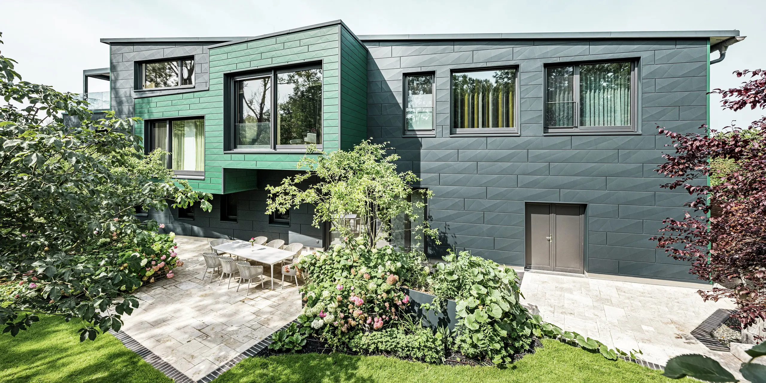 Das Einfamilienhaus in Türkheim beeindruckt durch seine außergewöhnliche Fassade, die mit PREFA Siding X-Paneelen in den Farben Anthrazit und Moosgrün verkleidet ist. Die klaren Linien und die markante Farbwahl der Aluminiumpaneele verleihen dem Gebäude eine moderne und elegante Ausstrahlung. Im Vordergrund ist der großzügige Gartenbereich mit einer stilvollen Terrasse und einem Esstisch mit modernen Stühlen zu sehen. Bunte Blumenbeete und gesunde Pflanzen sorgen für einen lebendigen, grünen Kontrast zur robusten Fassade. Die langlebige und pflegeleichte Aluminiumfassade schützt das Gebäude und sorgt für zeitlose Schönheit.
