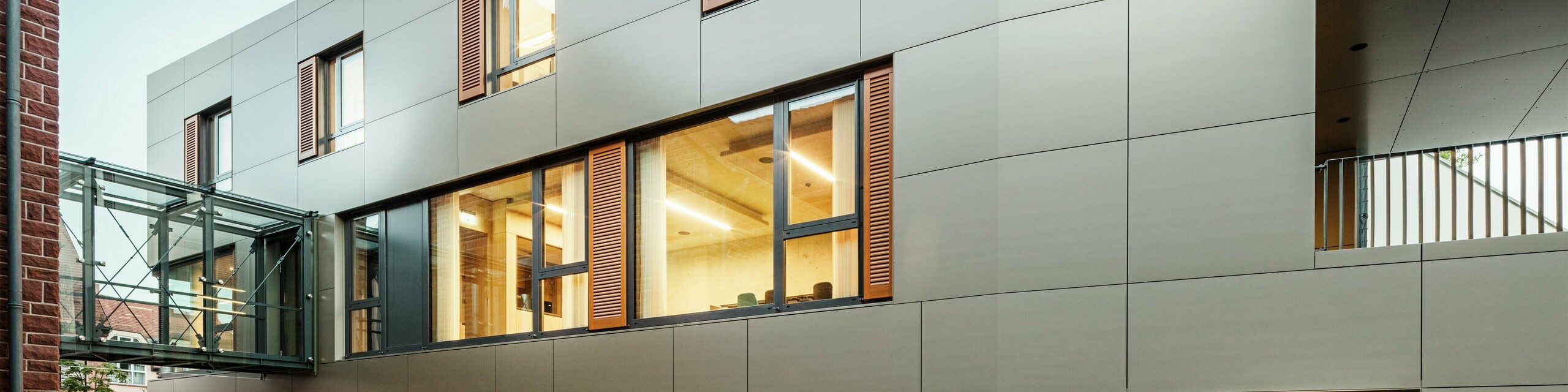 Moderne Fassade am Rathaus in Alzenau in Deutschland mit PREFA Aluminium Verbundplatten in Bronze, kombiniert mit Fensterläden und begrünten Pflanzflächen. Die hochwertige Aluminiumverkleidung sorgt für ein elegantes, langlebiges Erscheinungsbild und ist ideal für nachhaltige Bauprojekte. Hier ist die Seite gut ersichtlich.