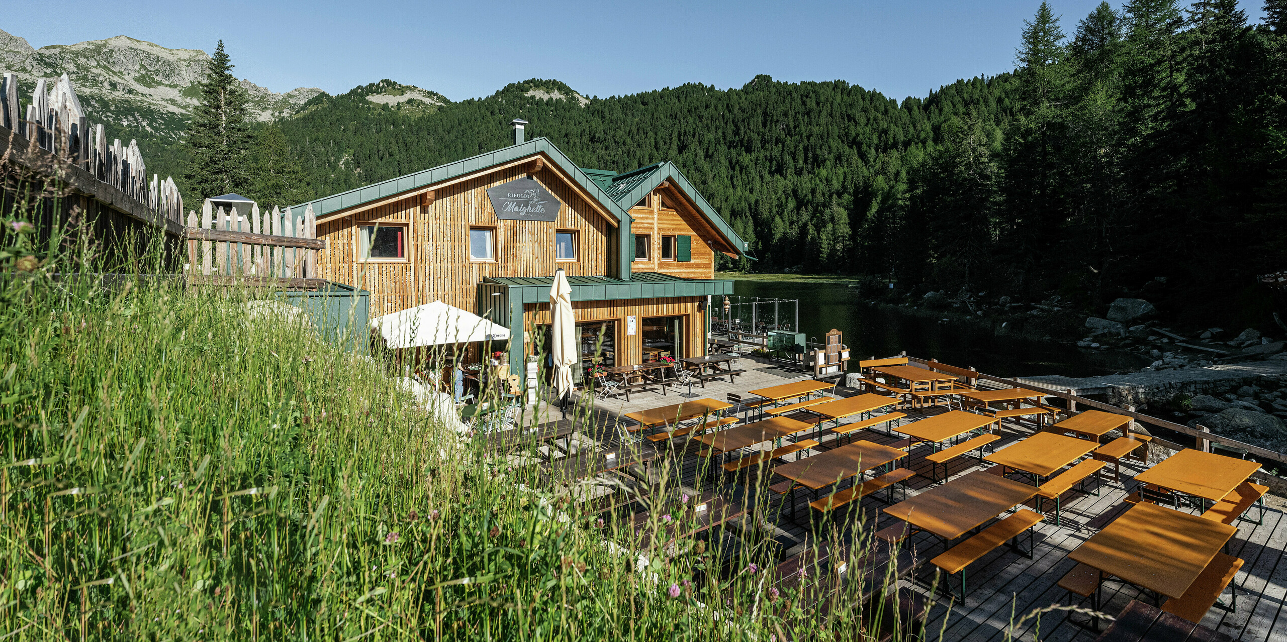 Das Rifugio Malghette mit PREFALZ-Dach in P.10 Moosgrün fügt sich harmonisch in die alpine Natur ein. Das wetterfeste Aluminiumdach schützt die Hütte zuverlässig auf 1.888 m Höhe. Die großzügige Sonnenterrasse direkt am See lädt Wanderer und Gäste zum Verweilen ein.