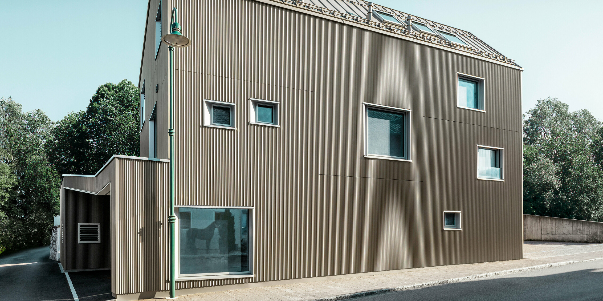 Blick von der Straße auf ein zweistöckiges Einfamilienhaus in Frankenburg, Österreich, gekennzeichnet durch ein PREFA Komplettsystem. Das Dach wurde mit PREFALZ in Bronze eingedeckt.. Die Fassade präsentiert sich in einer subtilen Textur, die durch die vertikalen Linien des Zackenprofils entsteht und ein harmonisches Schattenspiel erzeugt. Das Haus besitzt eine asymmetrische Fensteranordnung, die den modernen und unkonventionellen Charakter des Designs betont. Der Anbau auf der linken Seite ist stilistisch an das Hauptgebäude angelehnt, wodurch ein stimmiges Gesamtbild entsteht. Die klaren, sauberen Linien der Architektur kontrastieren wirkungsvoll mit dem blauen Himmel und den Bäumen im Hintergrund.