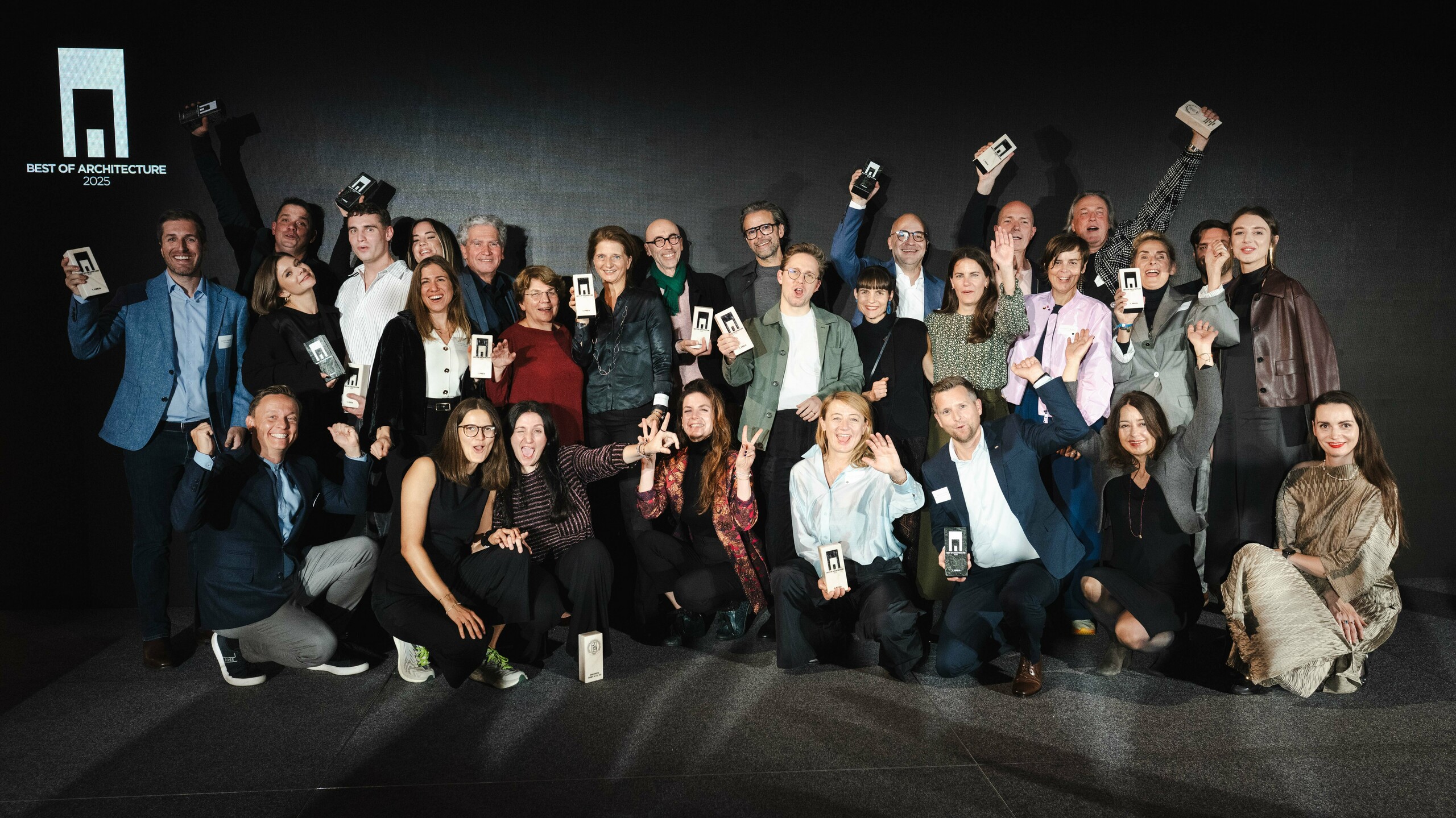 Gruppenfoto der Gewinner bei der Preisverleihung ‚Wohnbauten des Jahres‘ 2025: Die Preisträger stehen vor einer eleganten schwarzen Bühne mit dem Logo ‚Best of Architecture‘ und halten stolz ihre Auszeichnungen in den Händen. Dynamische Pose, moderne Kleidung und ein stimmungsvolles Lichtdesign unterstreichen die Bedeutung des Events. Die Szene symbolisiert Innovation und Architekturqualität.