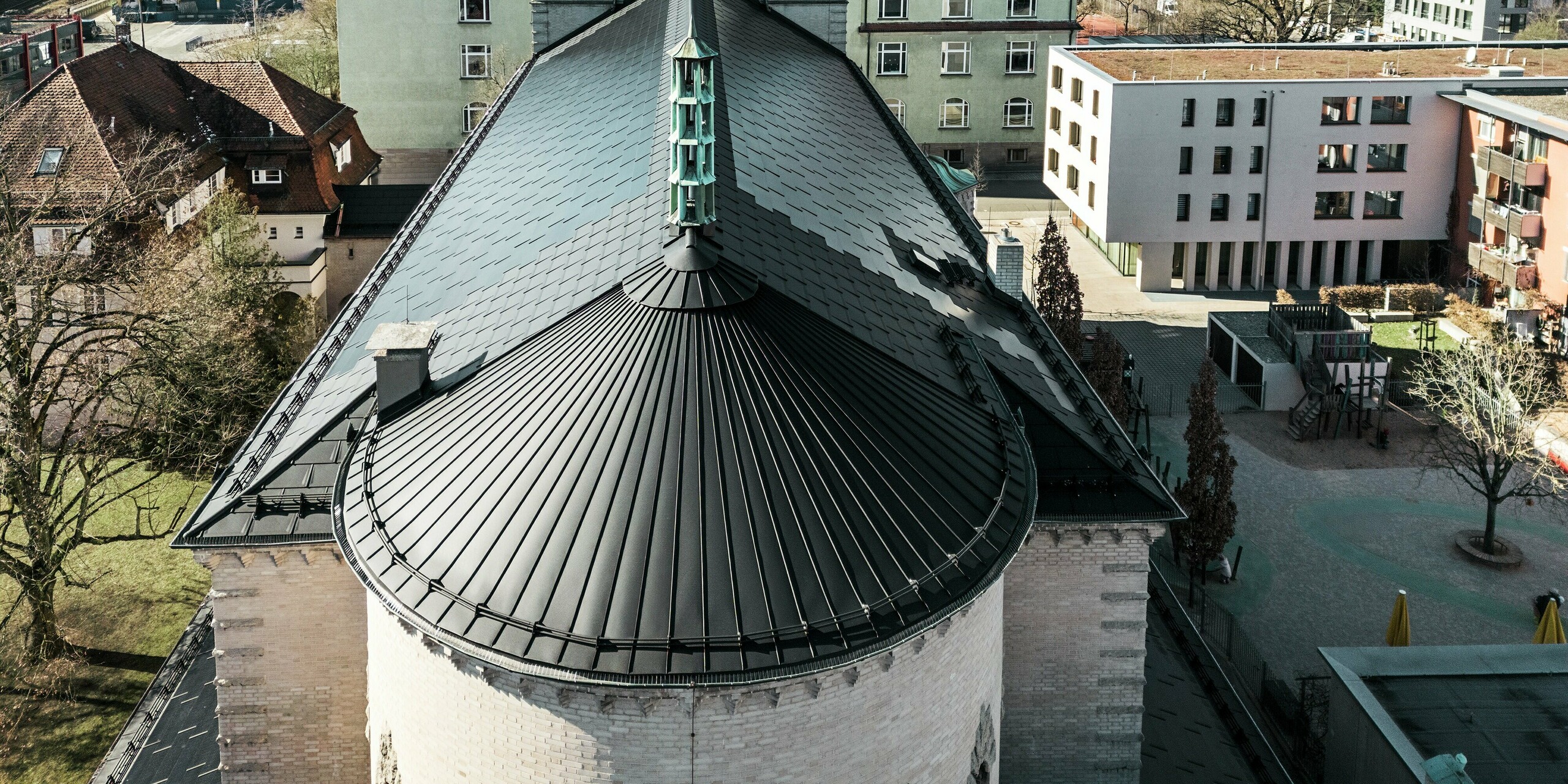Das Bild zeigt das markante, rund gewölbte Dach auf der Rückseite der St. Karl Borromäus Kirche in Nürnberg. Die Gebäuderundung wurde mit einer Stehfalzdeckung aus schwarzem Aluminium eingedeckt. Das Satteldach, das den Hauptteil des Gebäudes schützt, wurde mit schwarzen PREFA Solardachplatten eingedeckt. Die innovativen Solardachplatten fungieren als optisches Highlight und liefern gleichzeitig Strom aus erneuerbarer Energie. Der Blick auf das Dach und die zentral positionierte Turmspitze verdeutlicht die harmonische Integration der modernen Solartechnologie in die historische Architektur. Im Hintergrund sind sowohl traditionelle als auch moderne Gebäude zu sehen, was die gelungene Verbindung von Denkmalschutz und nachhaltigem Bauen unterstreicht.
