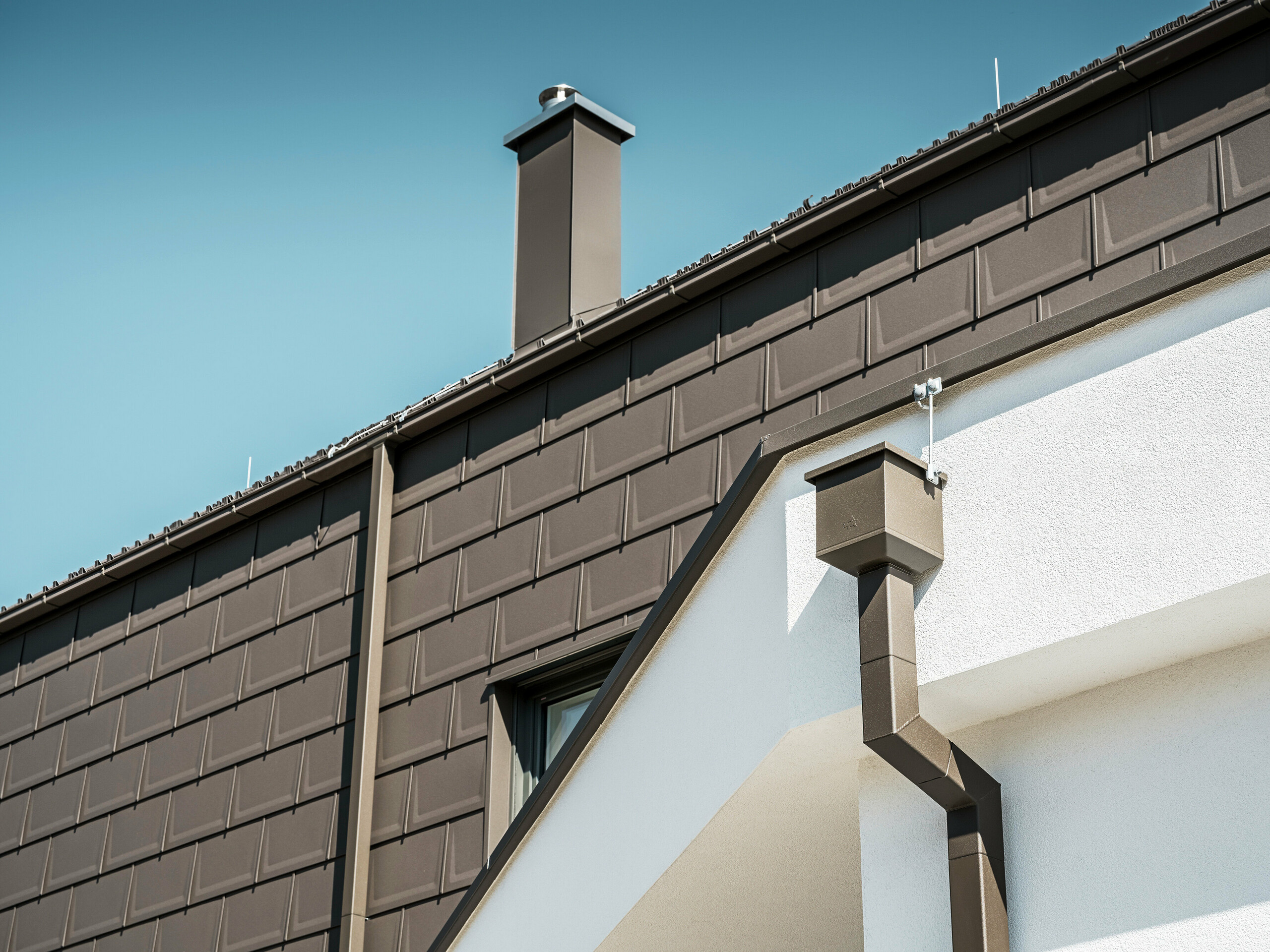Detailaufnahme des Dachrandes bzw. der Fassade eines Hauses in Neukirchen, Österreich, ausgestattet mit PREFA R.16 Dachplatten in P.10 braun. Sowohl die Dachplatten, die bei diesem Gebäude als Dach- und Fassadenverkleidung dienen, als auch die Dachentwässerung, bestehend aus Vierkantrohr, Wassersammelkasten und Kastenrinne, sind in der gleichen Farbe gehalten und bieten eine einheitliche, hochwertige Ausführung. Die Verwendung von PREFA Produkten verleiht dem Gebäude ein zeitgemäßes Aussehen, während die ästhetische Einbindung der Entwässerungselemente die funktionale Eleganz des Designs unterstreicht.