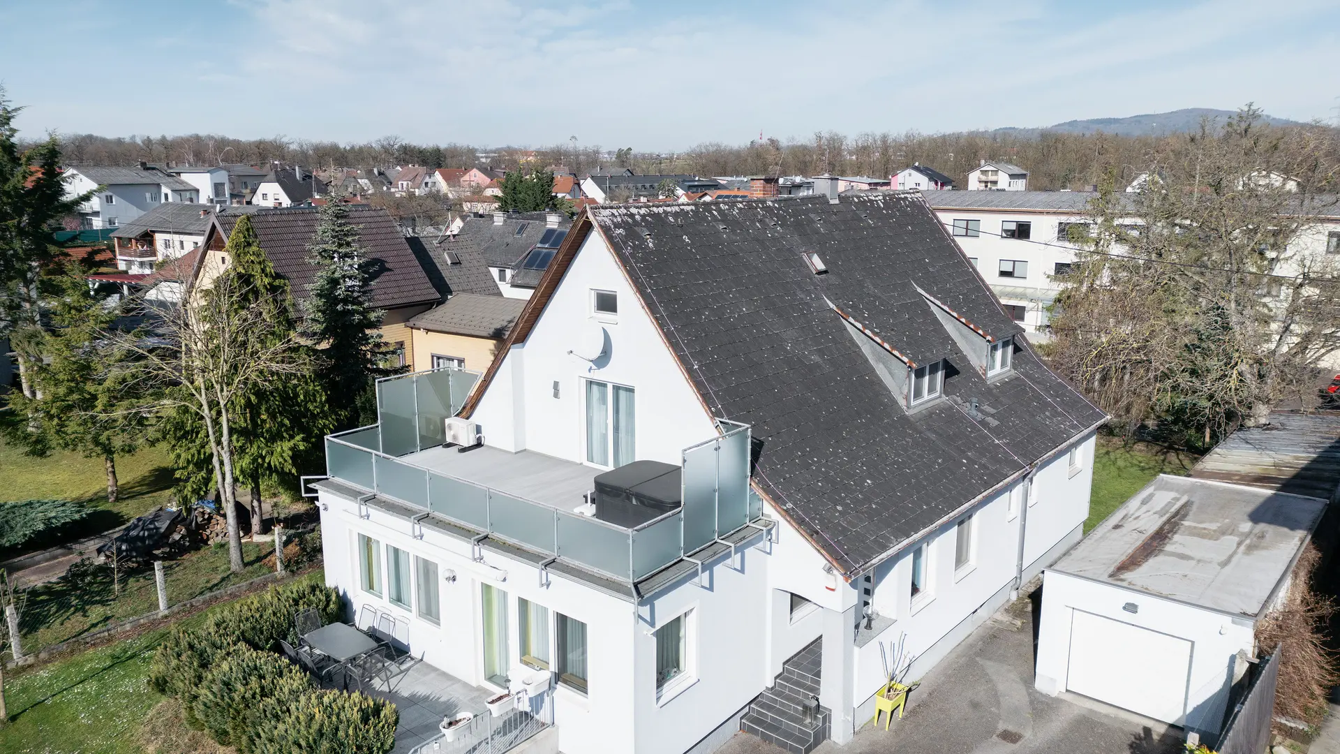 Vogelperspektive auf ein Gebäude mit einem alten Eternit-Dach, das von Schmutz und Flechten bedeckt ist. Auf dem Dach sind keine Solaranlagen sichtbar. Der Balkon im Vordergrund hat Glasgeländer und befindet sich auf der oberen Etage des Gebäudes.