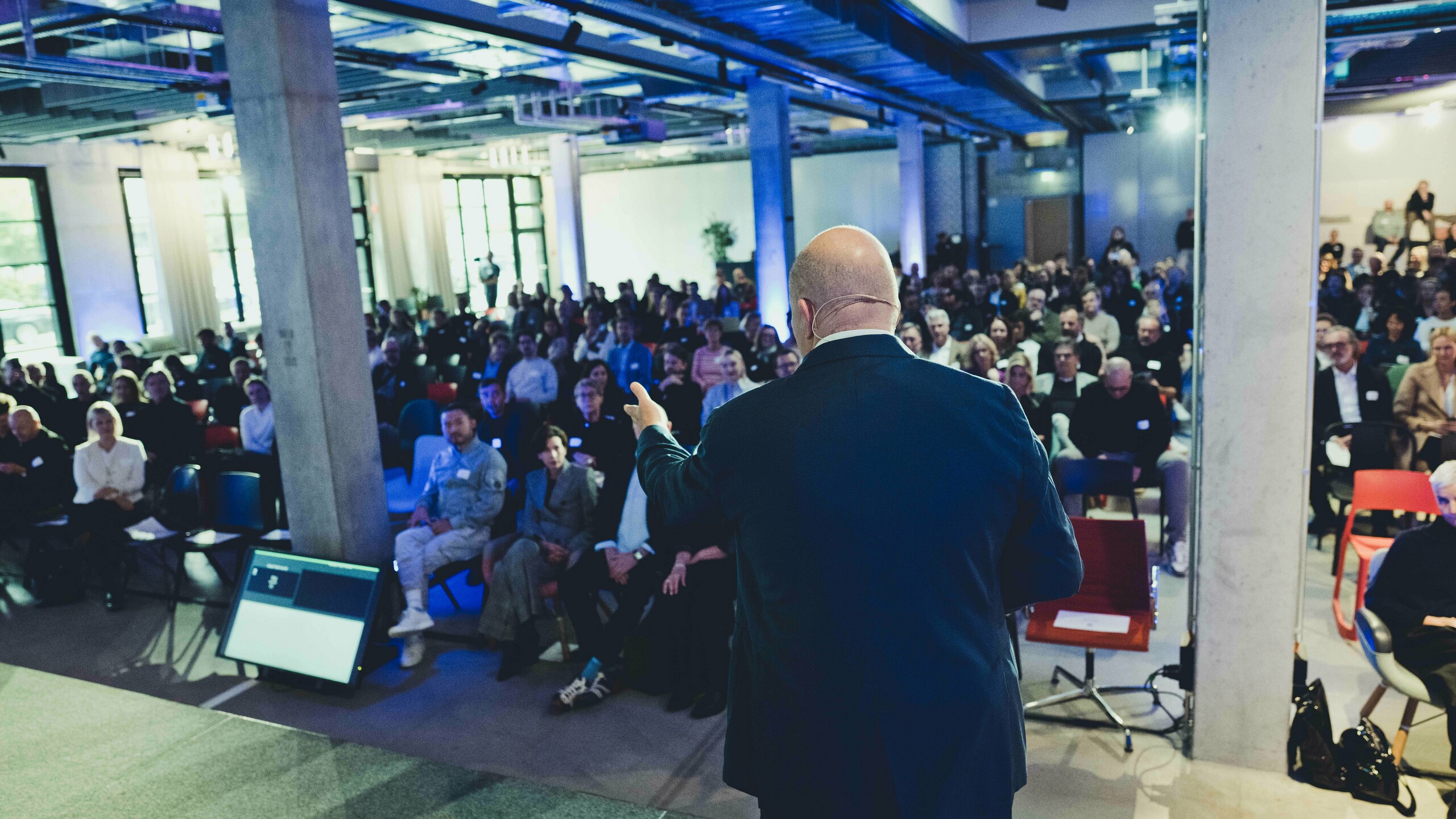 Spannende Vortrags-Szene bei der Preisverleihung ‚Wohnbauten des Jahres‘ 2025: Blick von der Bühne auf ein voll besetztes Auditorium in modernem, lichtdurchflutetem Raum mit industriellem Design. Im Vordergrund steht der Redner vor einer großen Leinwand, während die Zuhörer aufmerksam dem Vortrag folgen. Die Veranstaltung unterstreicht Innovation und Architekturtrends.