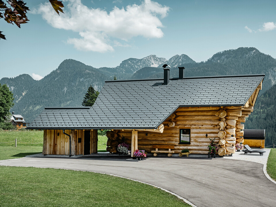 Blockhaus in Gosau, Österreich, mit einem PREFA Dachsystem in der Farbe Anthrazit. Die bewährten Dachplatten R.16 schaffen einen ansprechenden Kontrast zum natürlichen Holz des Blockhauses. Im Hintergrund erstreckt sich ein majestätisches Bergpanorama, das die ruhige und malerische Lage des Hauses unterstreicht. Die detailreiche Aufnahme fängt die Symbiose aus traditioneller Baukunst aus Österreich und moderner Dachtechnologie ein und zeigt die robuste und ästhetische Qualität der PREFA Aluminiumprodukte.